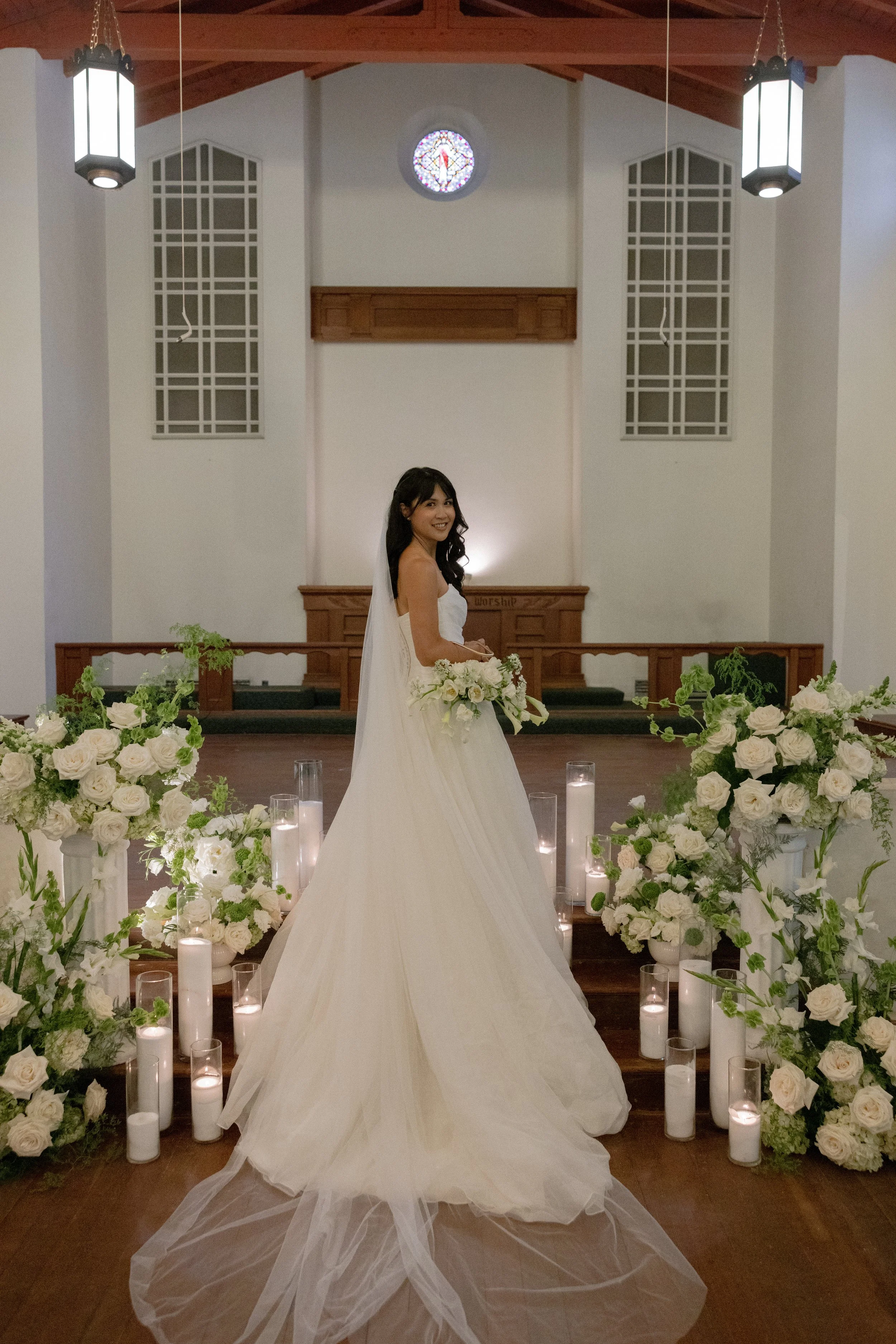 A smiling bride standing in a church decorated with white roses and candles, wearing a white wedding gown with a veil, holding a bouquet of white flowers. The church interior has high ceilings, wooden beams, stained glass window, and hanging lantern 