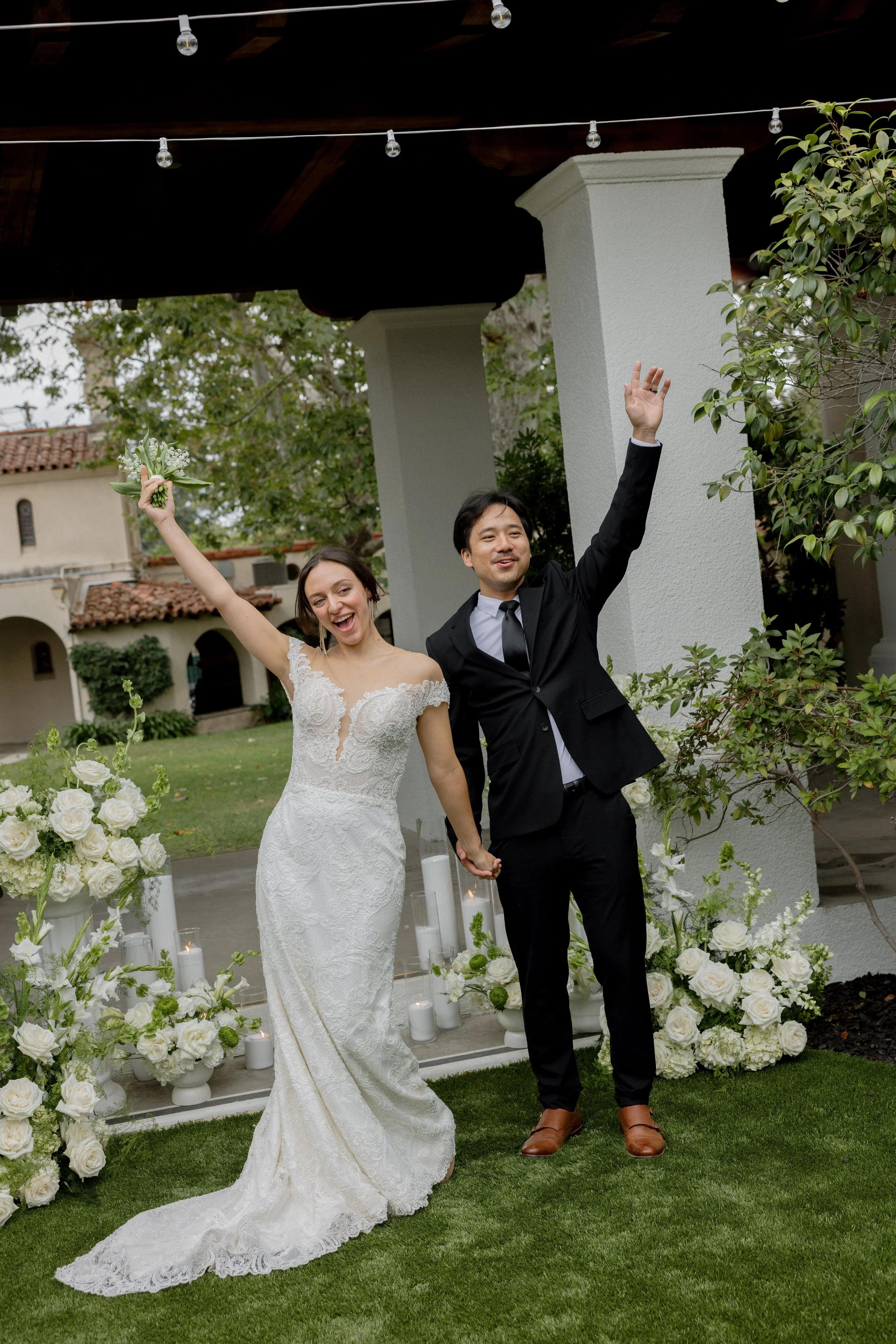 A bride and groom, holding hands, celebrating their wedding outdoors. The bride is in a white lace wedding gown, and the groom is in a black suit with a tie. The scene is decorated with white flowers and candles, with a white column and greenery in t