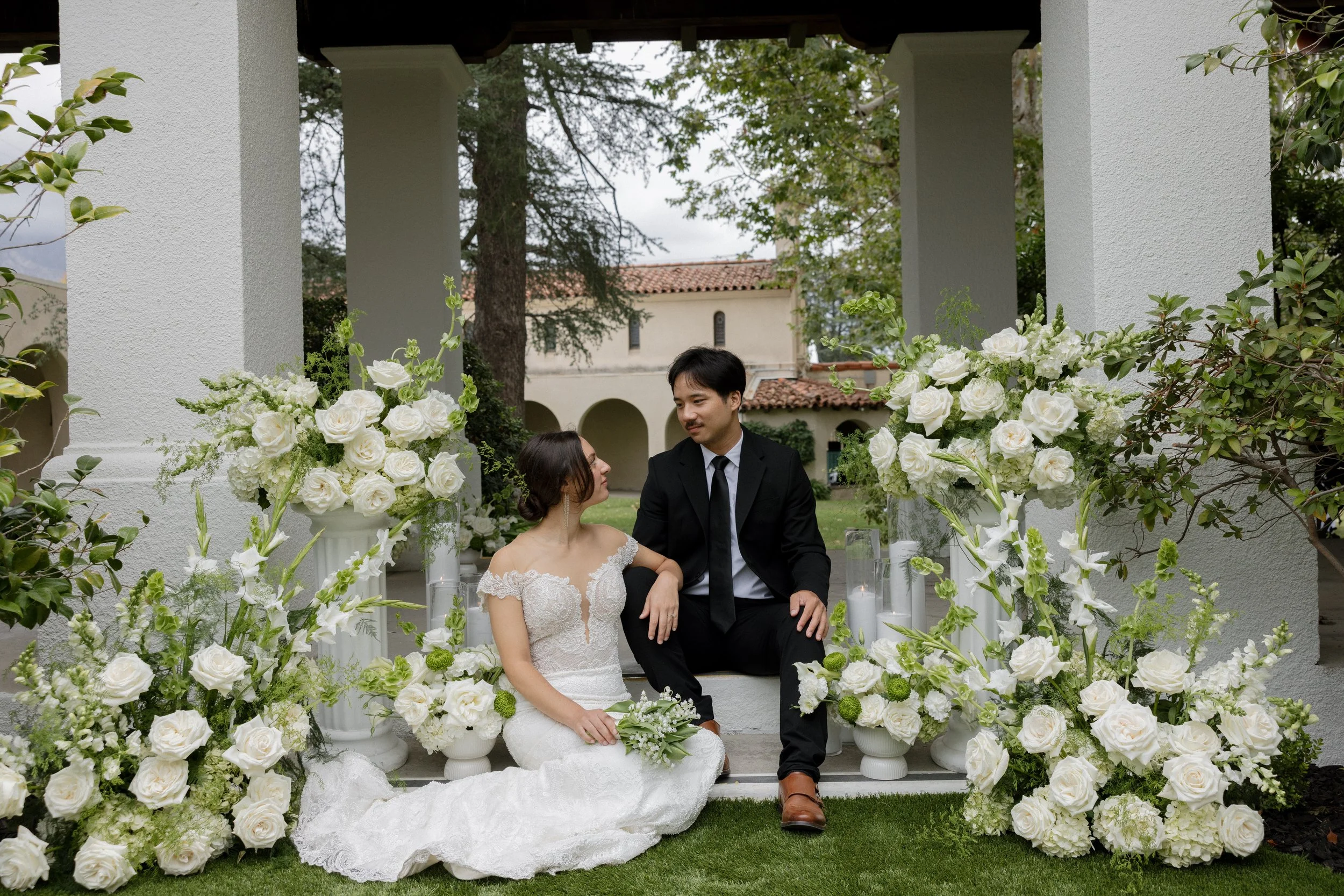 A bride and groom sitting on a white bench surrounded by white floral arrangements in an outdoor wedding setting.