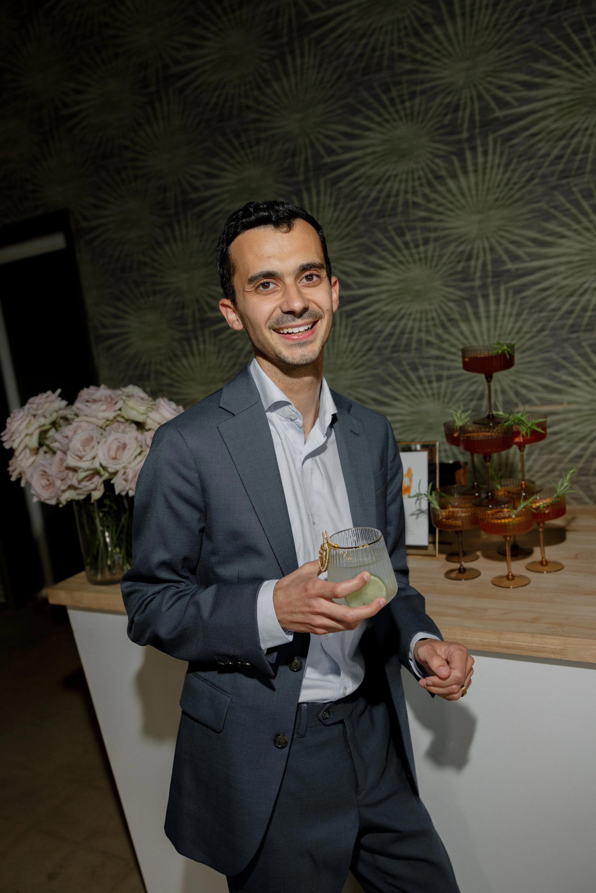 A man in a gray suit holding a cocktail glass at a social event, standing near a table with floral decorations and a tiered tray of desserts, against a patterned wall background.