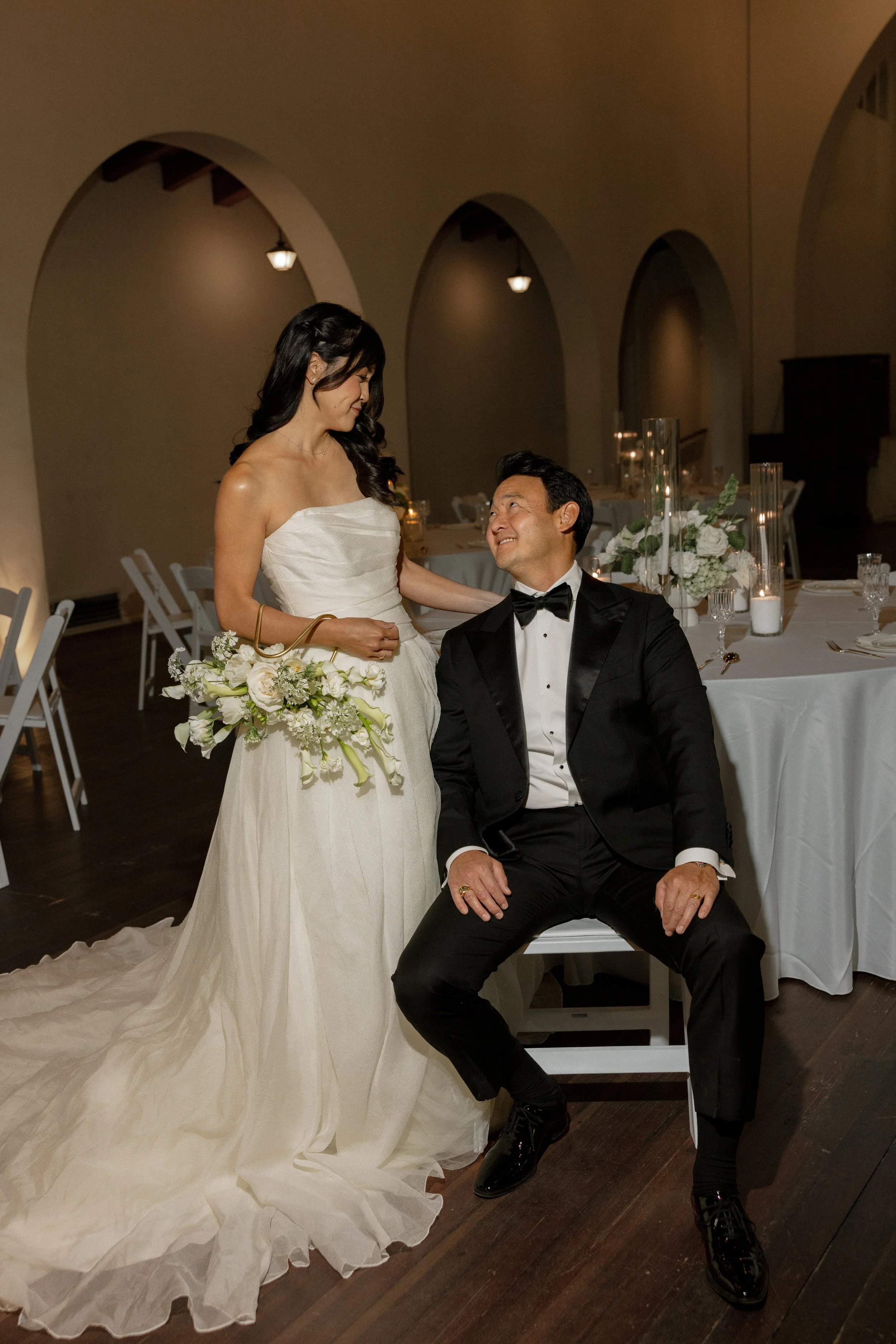 A bride in a strapless white wedding gown holding a bouquet of white flowers, looking at a seated groom in a black tuxedo with a bow tie, in a decorated wedding reception hall.