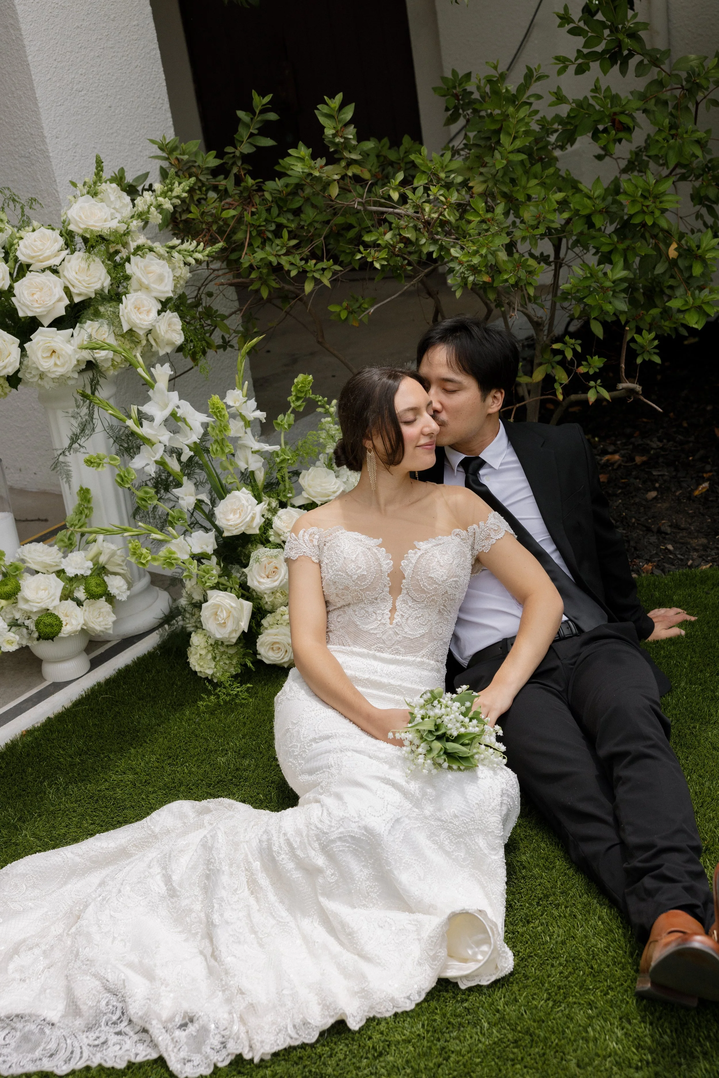 A bride and groom sitting outdoors on green grass, surrounded by white flowers and greenery. The groom is giving the bride a gentle kiss on the forehead, and they both look peaceful with their eyes closed. The bride is wearing a white lace wedding dr