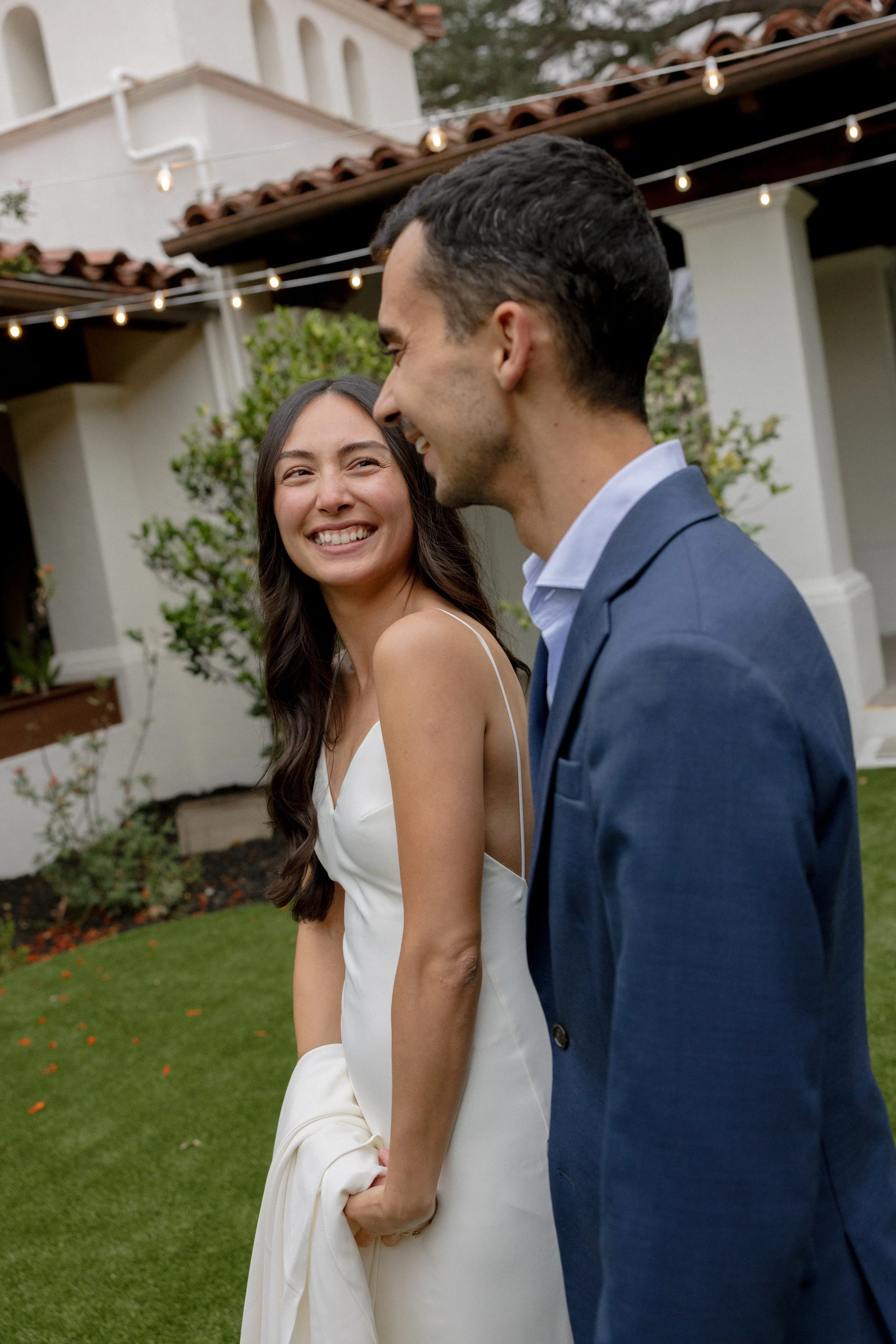 A smiling woman and man dressed in wedding attire are outdoors at sunset, standing close together with greenery in the background.