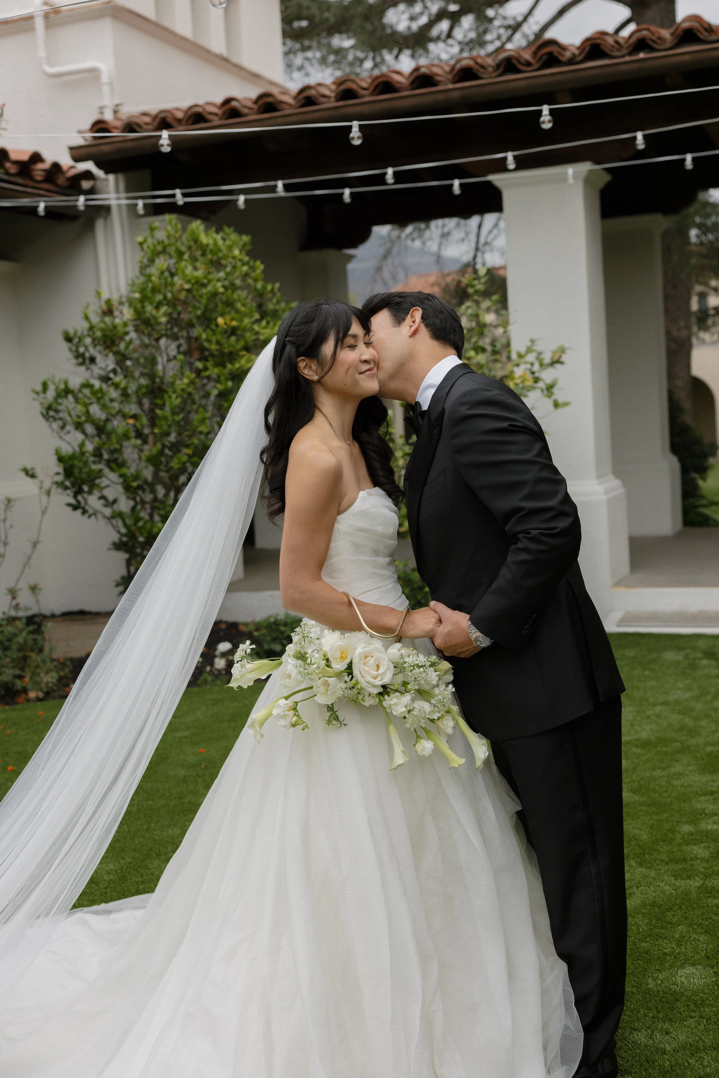 A bride and groom sharing an intimate moment outdoors, the bride in a strapless wedding gown holding a bouquet of white flowers, the groom in a black tuxedo, in front of a white building with a red tile roof, string lights overhead, and greenery in t