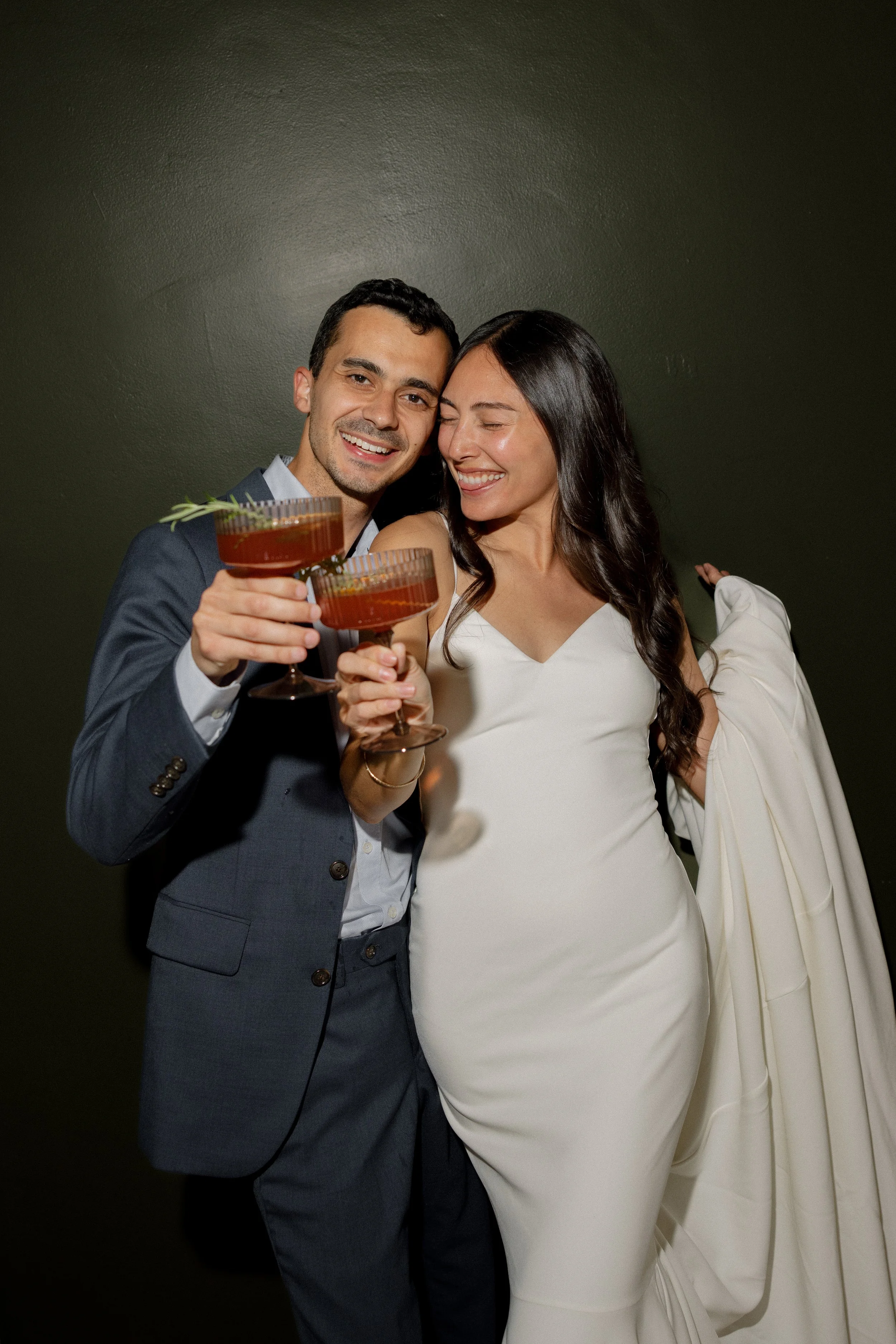A man and woman smiling and celebrating with drinks, dressed in formal attire, against a dark background.
