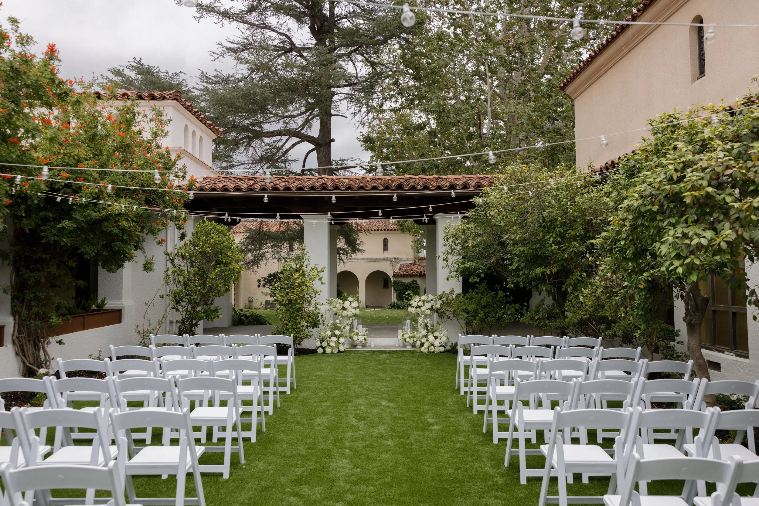 Outdoor wedding setup with white chairs arranged on green grass facing an altar with white flowers, surrounded by trees and buildings with Spanish-style red tile roofs, decorated with string lights.