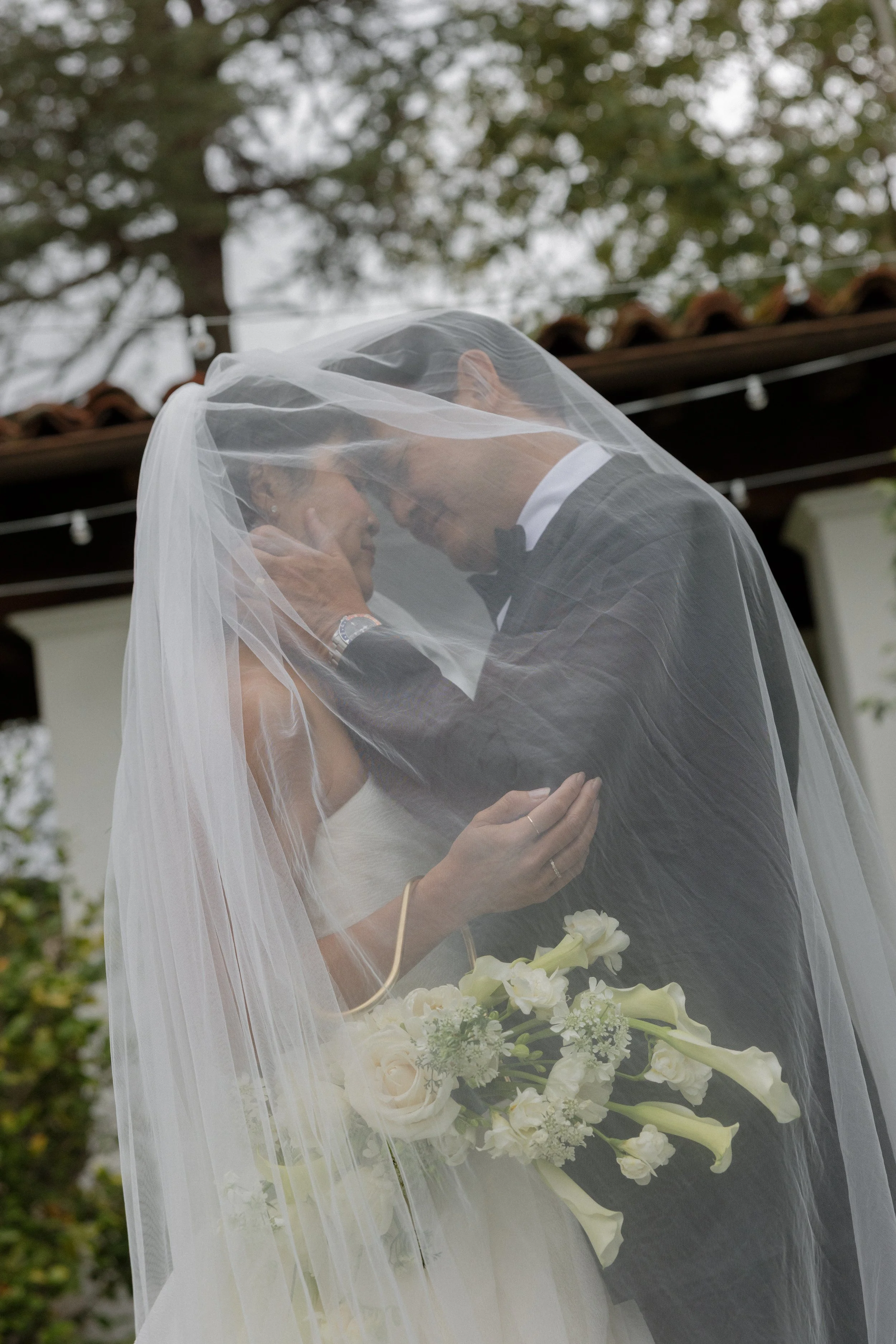 A bride and groom sharing a kiss under a wedding veil during an outdoor wedding ceremony, with trees and string lights in the background.