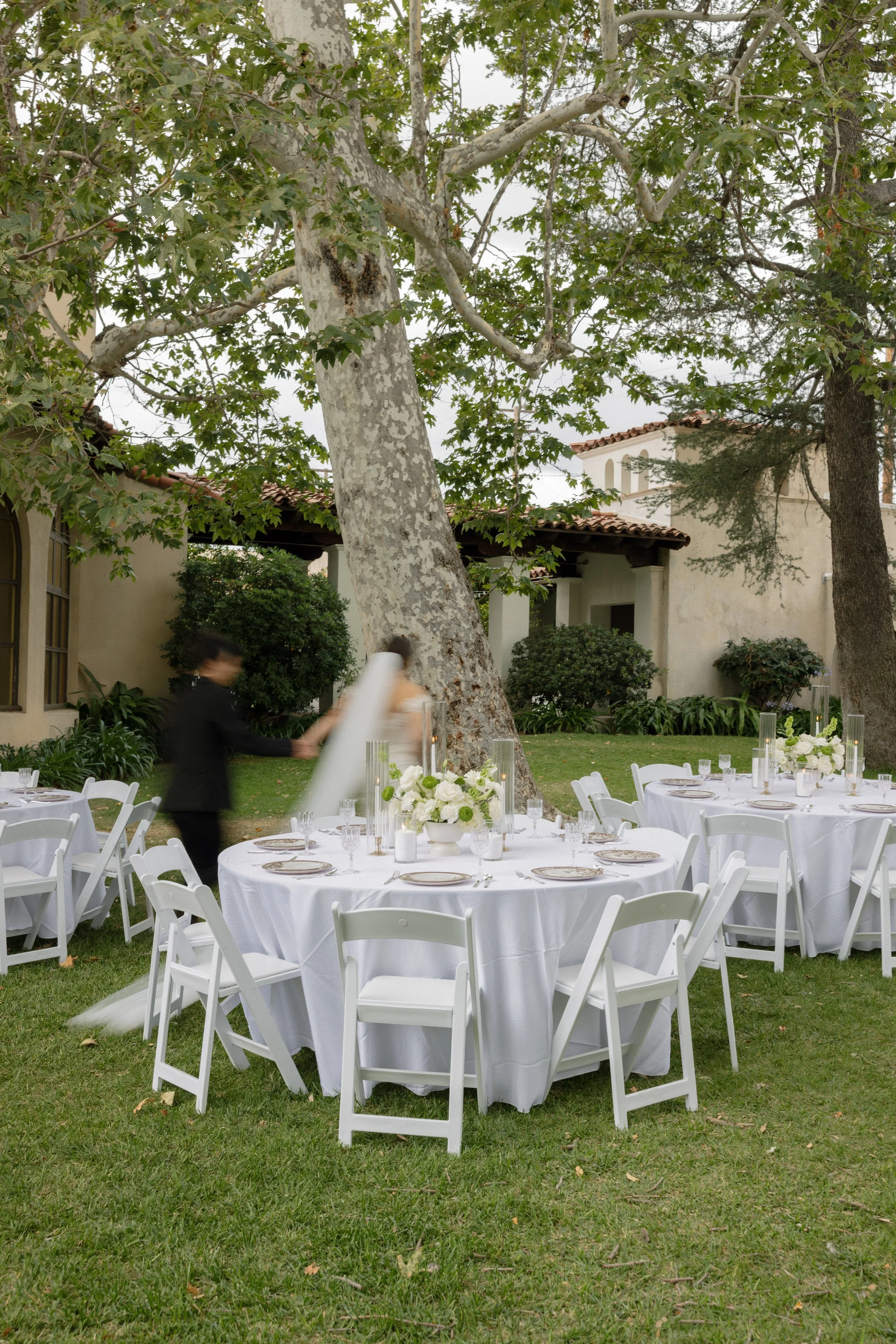 Outdoor event setup with round tables covered in white tablecloths, decorated with floral centerpieces and glassware, surrounded by white chairs on a grassy area. In the background, there are tall trees, some of which have peeling bark, and a building with a red-tiled roof and Mediterranean-style architecture. Two blurred people are moving near the tables.