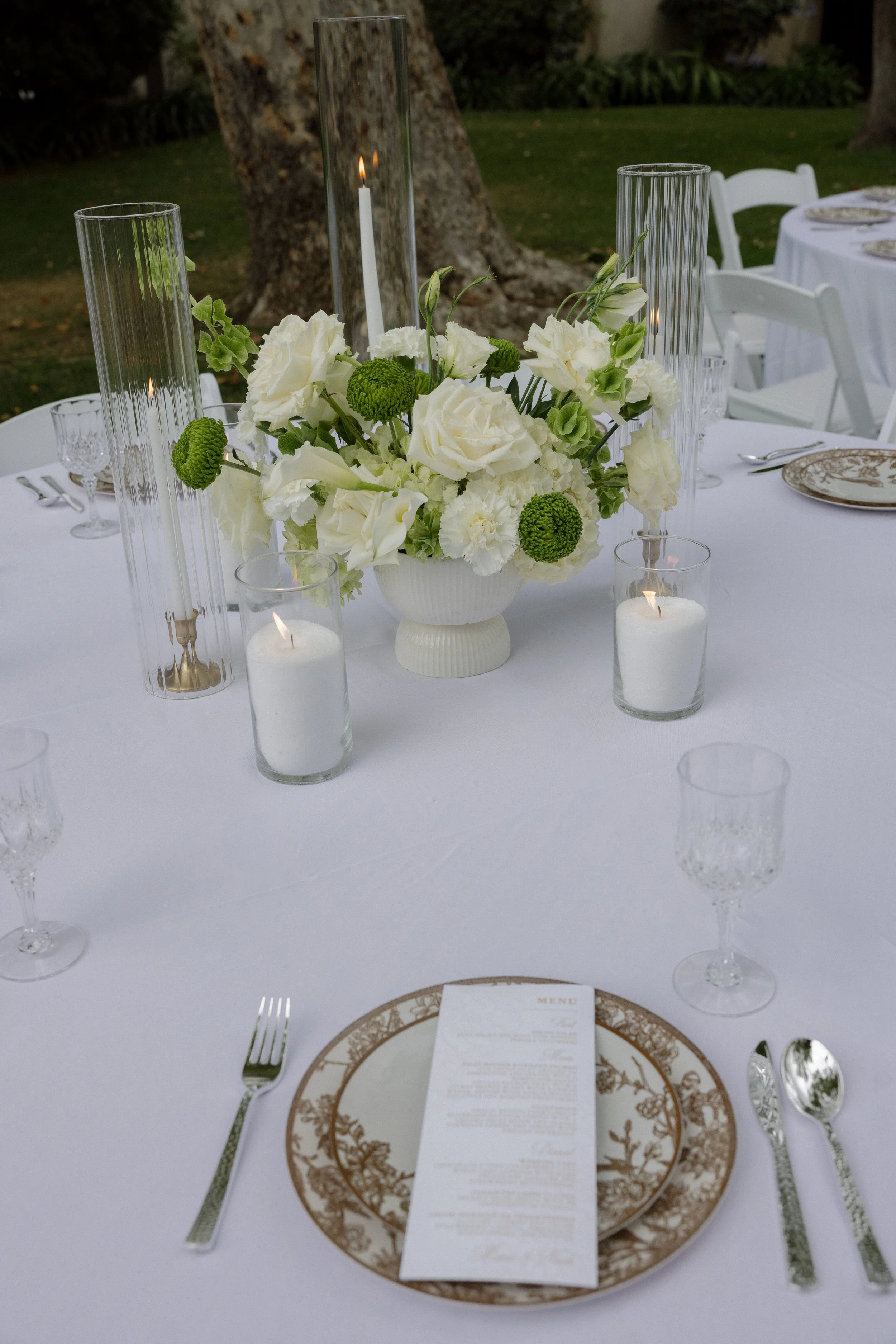 A dining table decorated with a white tablecloth, a flower centerpiece with white and green flowers, tall glass candle holders with lit candles, and a place setting with a decorative plate, menu, silverware, and glasses. The setting appears to be out
