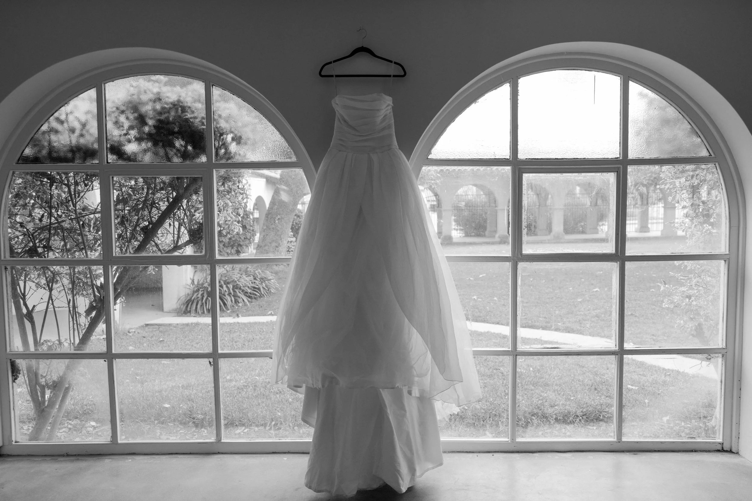 A wedding dress hanging on a hanger in front of large arched windows with a view of a garden outside.