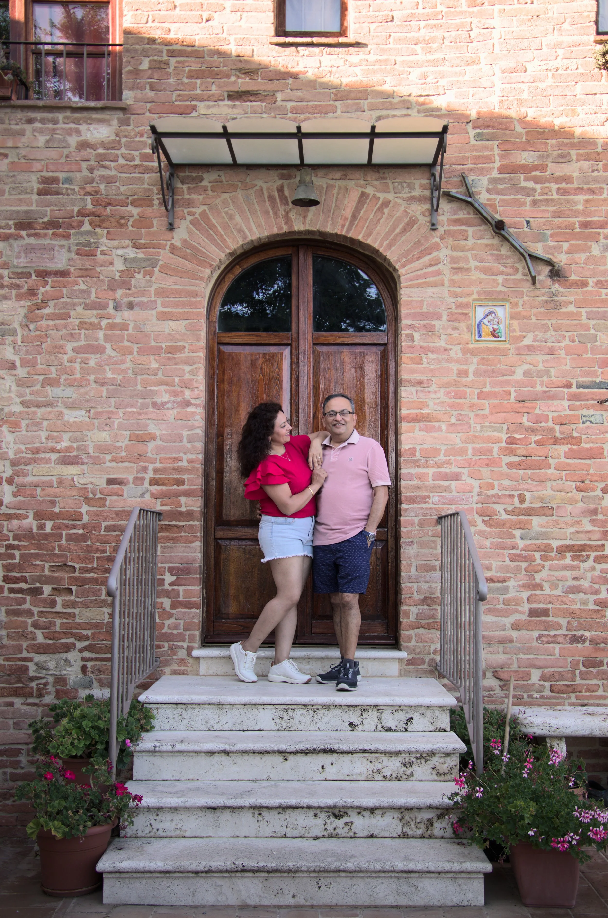 A man and woman standing on a stone staircase in front of a wooden door, smiling and looking at each other unposed and intimate