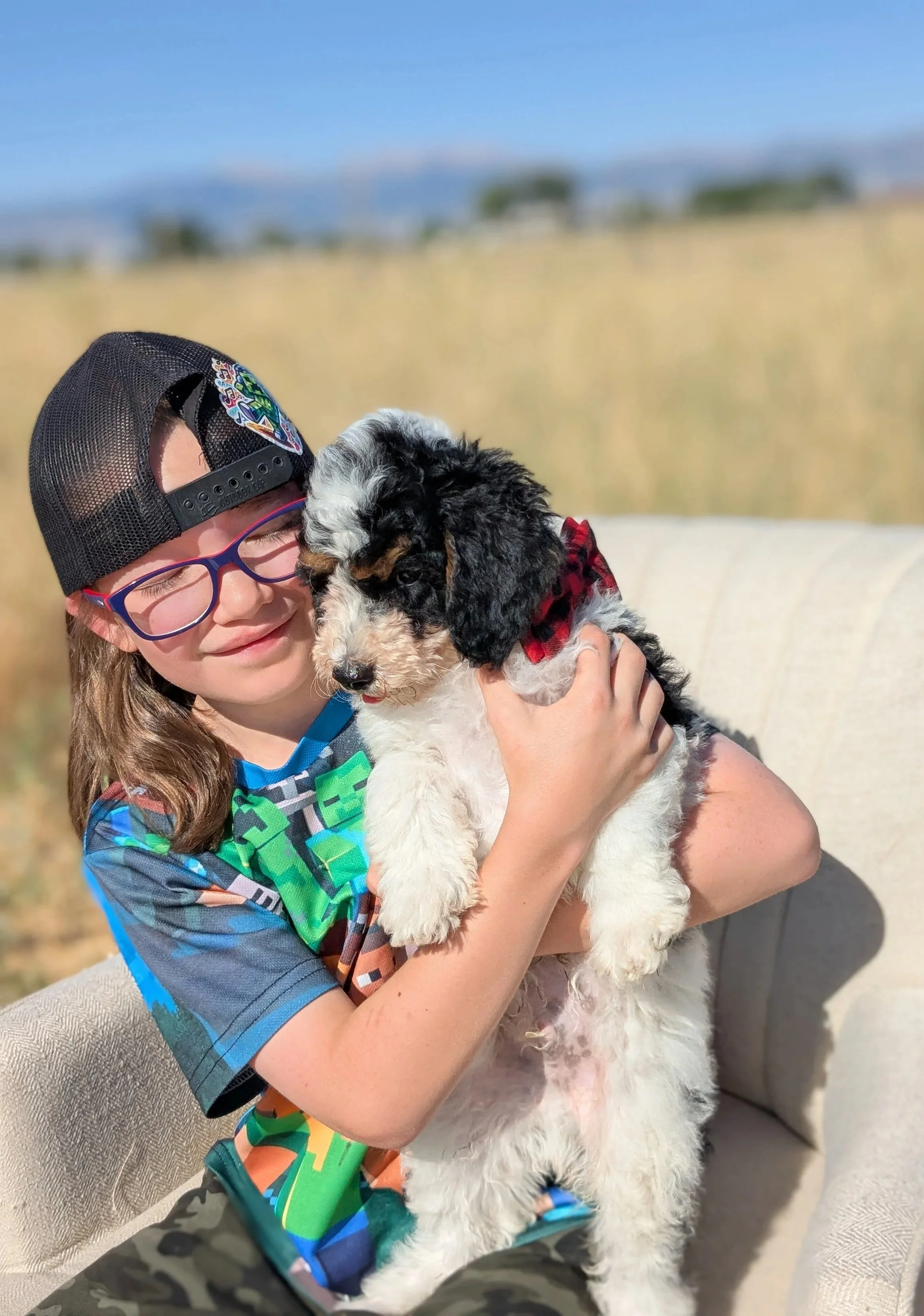 A young boy wearing a black cap backwards, red glasses, and a colorful shirt, is happily hugging a black and white puppy with floppy ears, outdoors with a blurred background of field and blue sky.