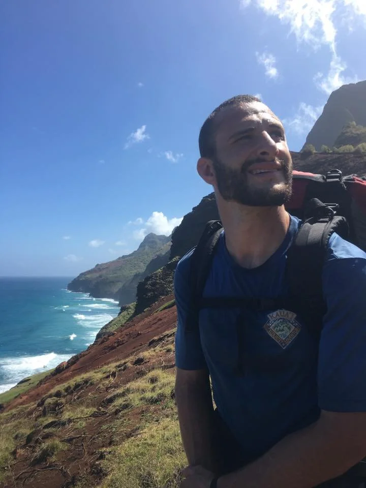 A man hiking along Kalalau trail with steep cliffs and the ocean on the left, smiling and looking to the side, carrying a backpack under a partly cloudy sky.