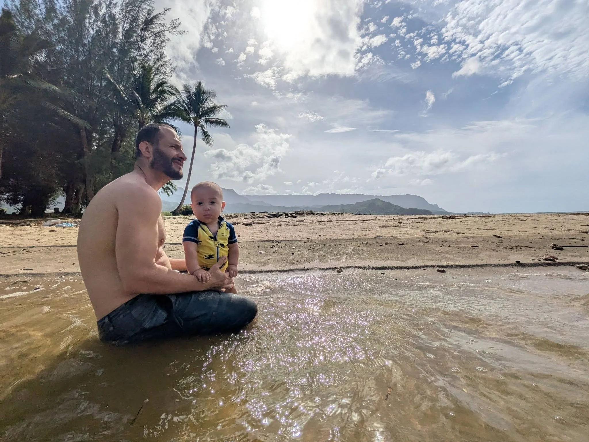A man and a young boy sitting in shallow ocean water near the beach with palm trees and mountains in the background under partly cloudy sky.