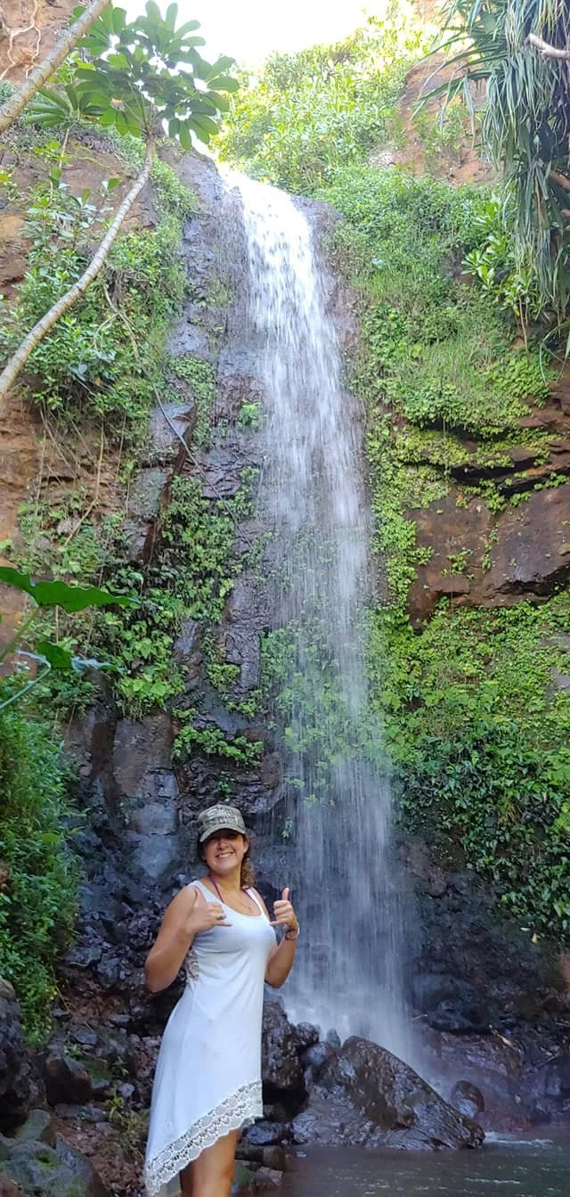 A woman in a white dress and camouflage hat standing at the base of a waterfall in a lush, green outdoor setting, throwing up shaka signs.