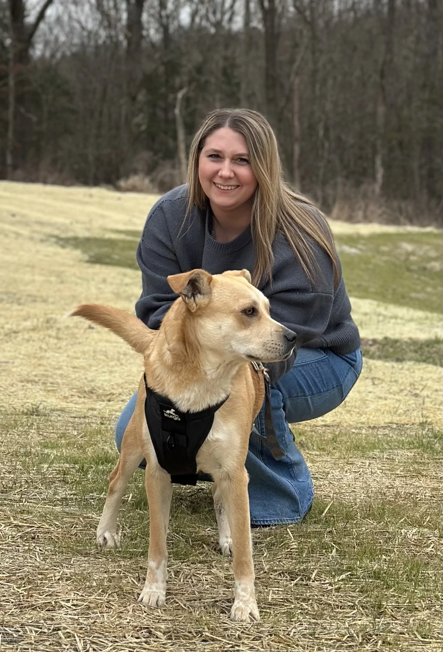 A woman with long blonde hair smiling and crouching on the ground outdoors with a yellow dog wearing a harness in front of her, set in a field with trees in the background.