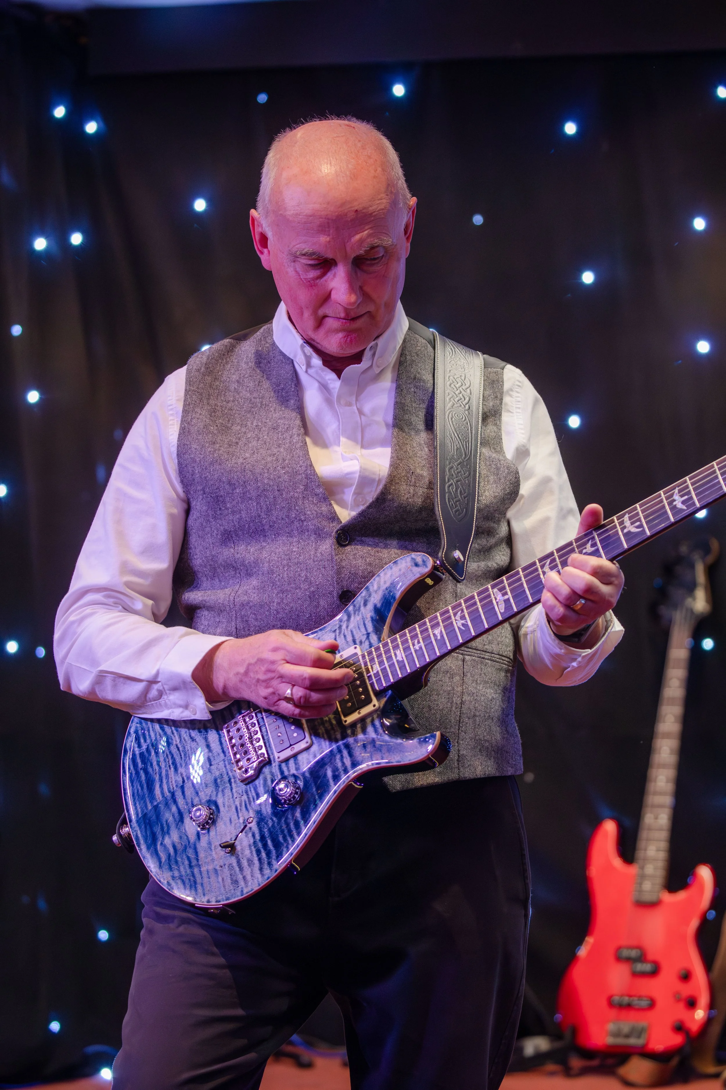 An elderly man playing an electric guitar on stage with a dark background featuring small bright lights and a red bass guitar in the background.