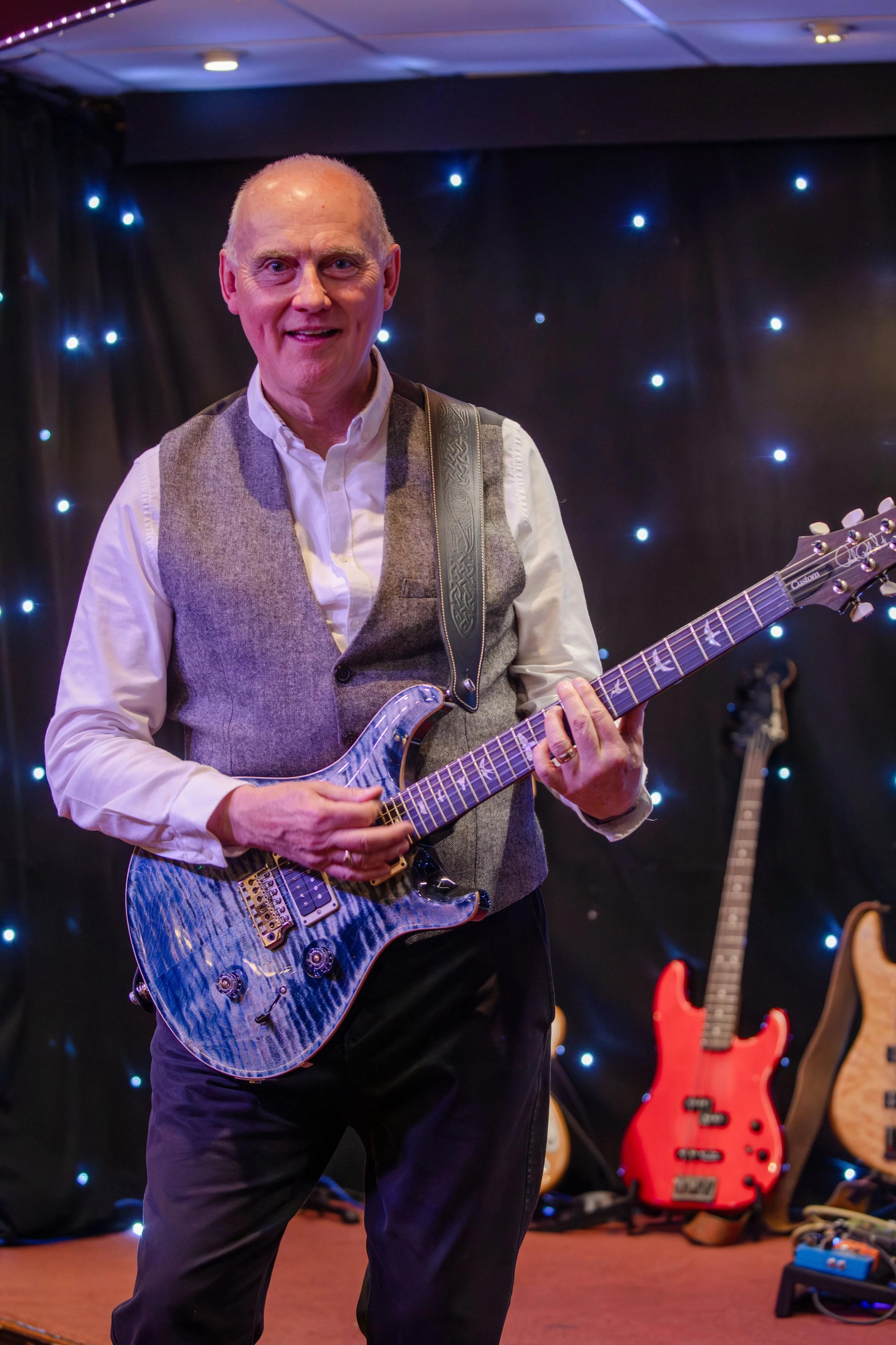 A smiling man playing an electric guitar on a stage decorated with blue fairy lights, with other guitars visible in the background.
