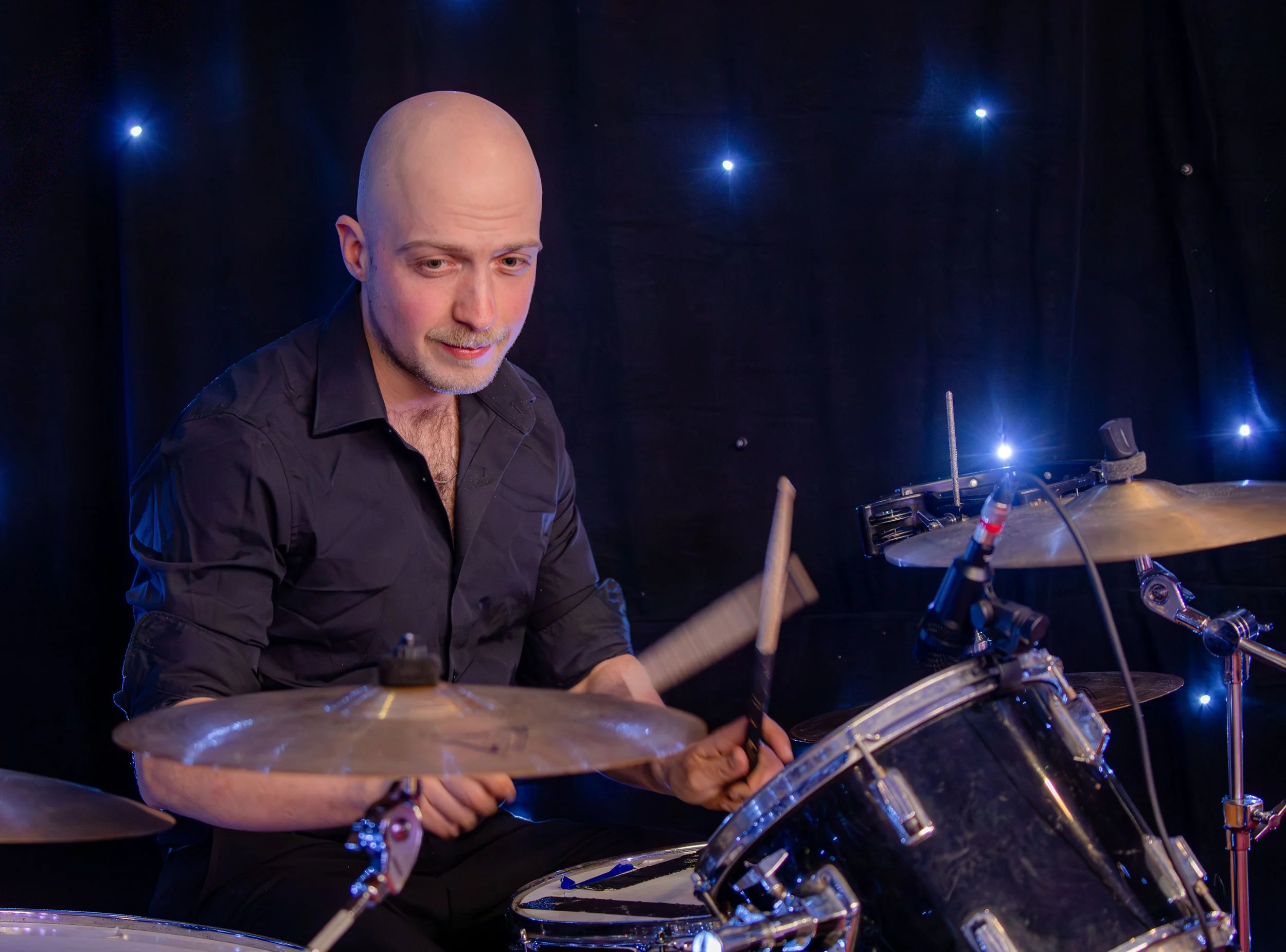 A bald man playing drums on stage with dark background and blue stage lights.