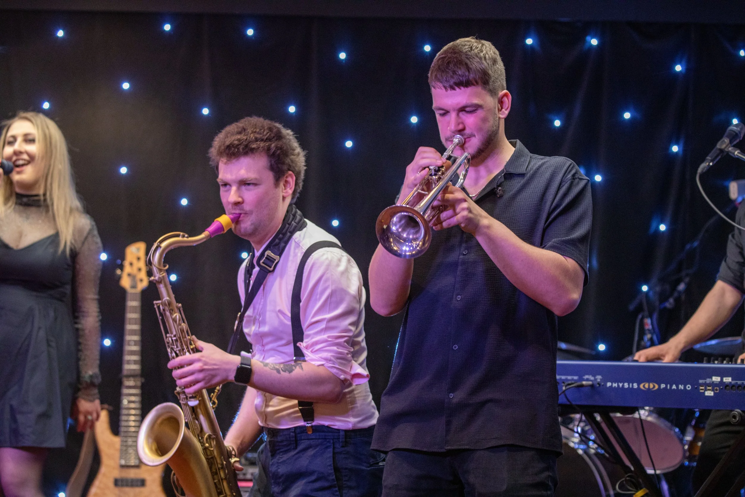 A group of musicians performing on stage with black background and blue star-like lights. A woman on the left sings into a microphone, with a need guitar visible beside her. Two men in the middle are playing the saxophone and trumpet, respectively. T