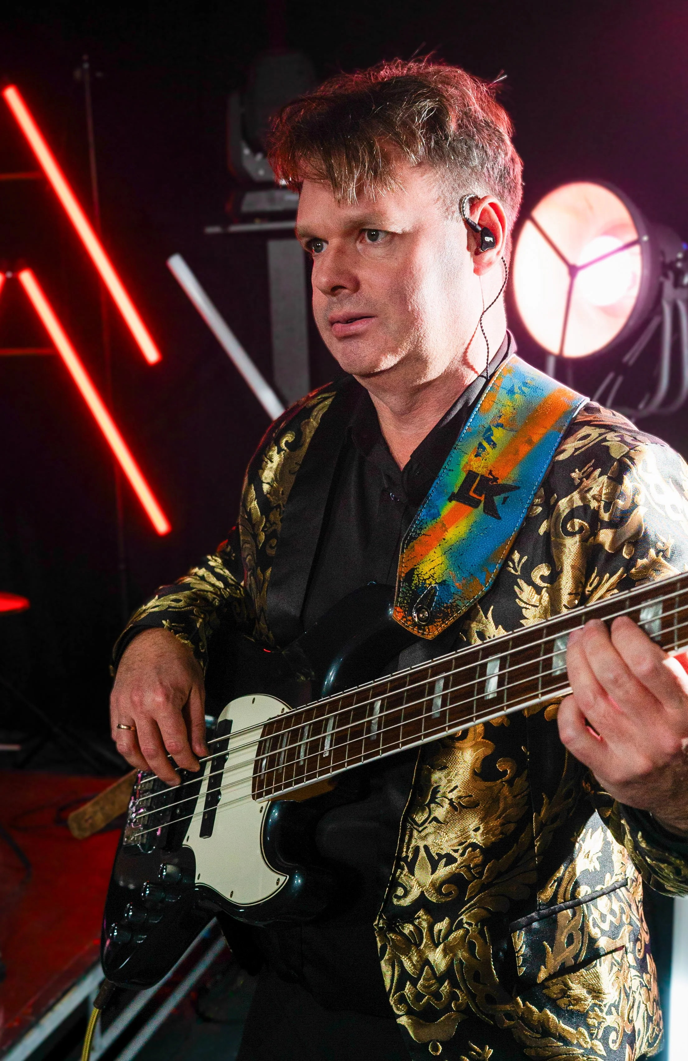 Man playing black and white electric guitar, wearing a black and gold patterned jacket and in-ear monitors, illuminated by stage lighting.
