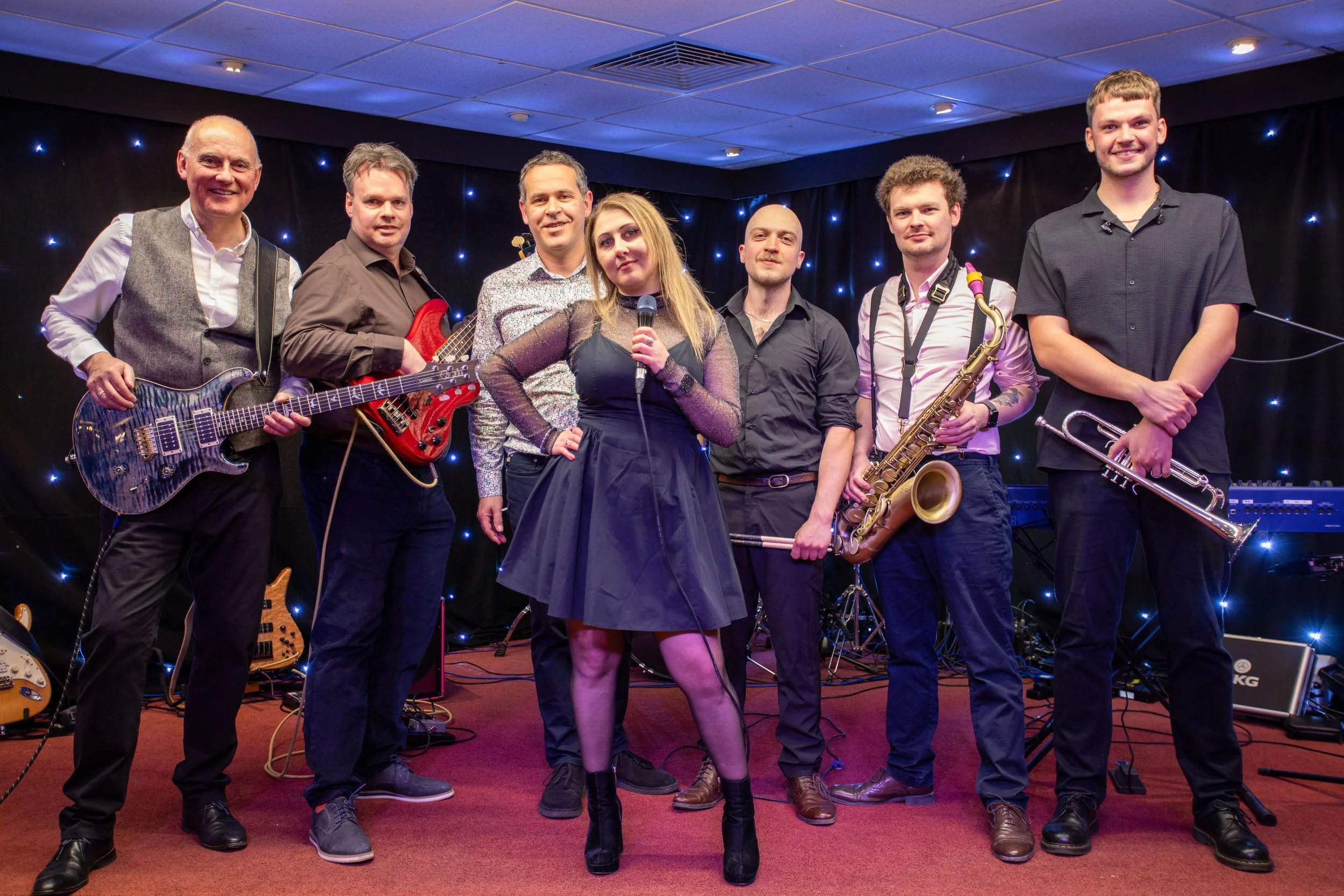 A music band performing on stage with seven members, including guitarists, a singer with a microphone, and a saxophonist. The background is decorated with small blue lights, and the stage has a red carpet.