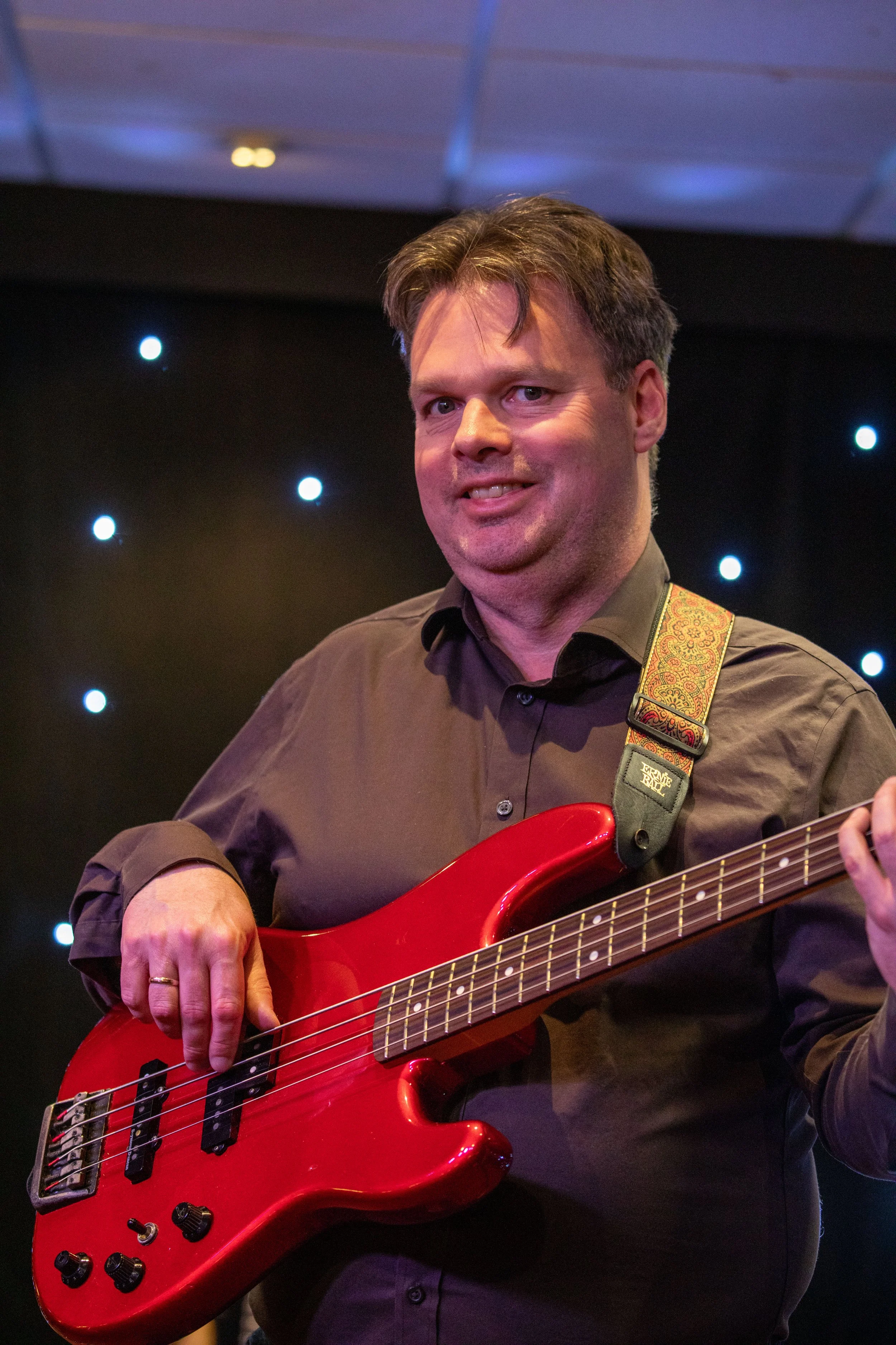A man playing a red electric bass guitar on a stage with black background and small bright lights.