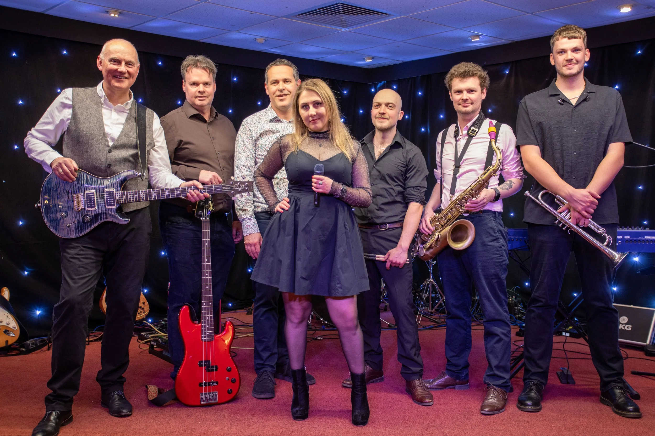 Re-Release wedding band - a group of seven musicians standing on a stage with black backdrop and star-like lights, holding musical instruments including a guitar, bass guitar, keyboard, and saxophone. A woman in the centre is holding a microphone.