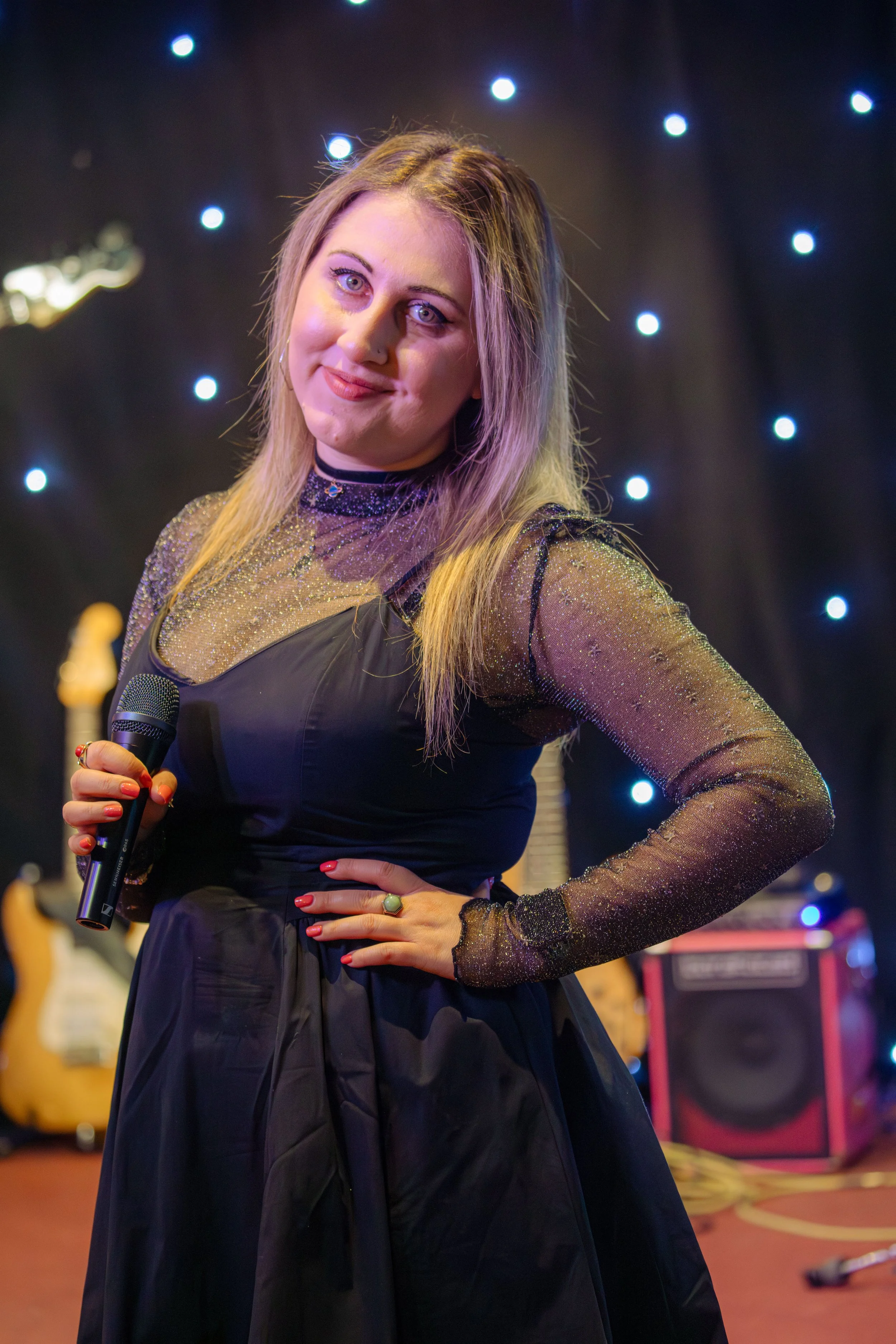 A woman with blonde hair, wearing a sparkly black dress and holding a microphone, posing in front of a black backdrop with small blue lights. There are musical instruments and equipment in the background.