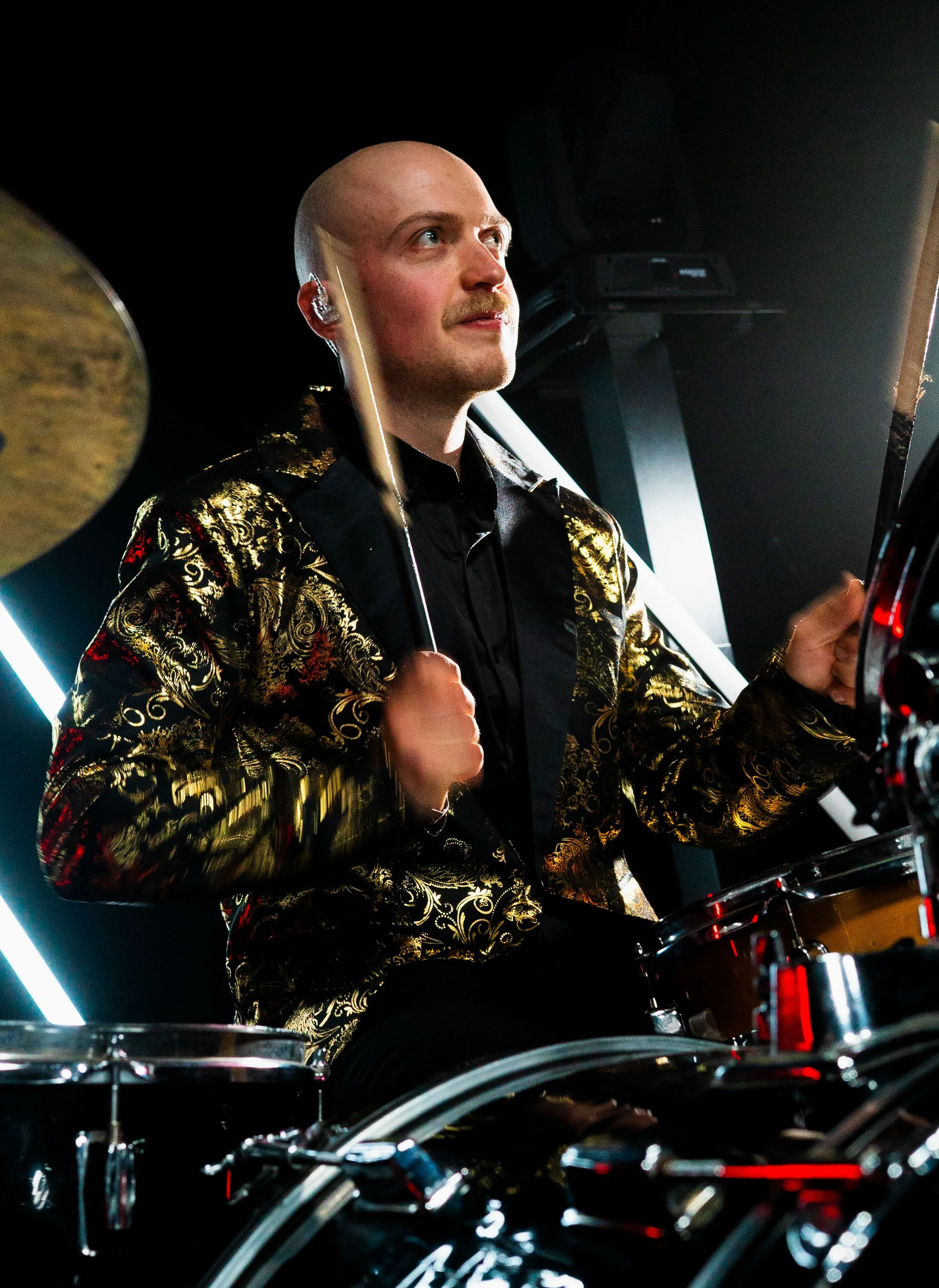 A man with a shaved head and mustache playing drums on stage, wearing a black and gold patterned jacket and in-ear monitors, with stage lighting in the background.