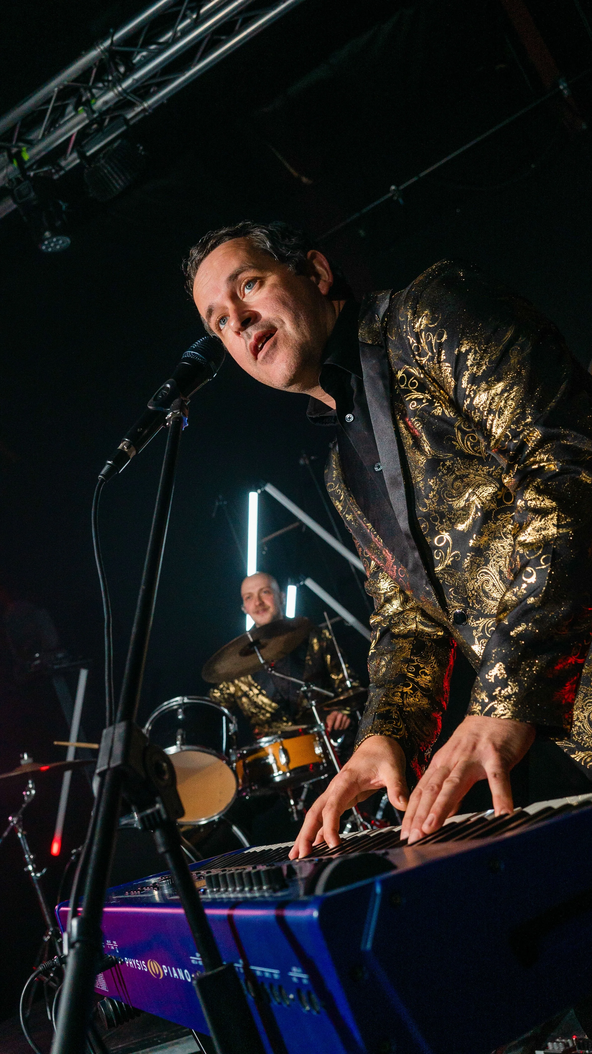 A man playing a keyboard and singing into a microphone on stage, with a drummer in the background, both wearing black and gold patterned outfits, under stage lighting.