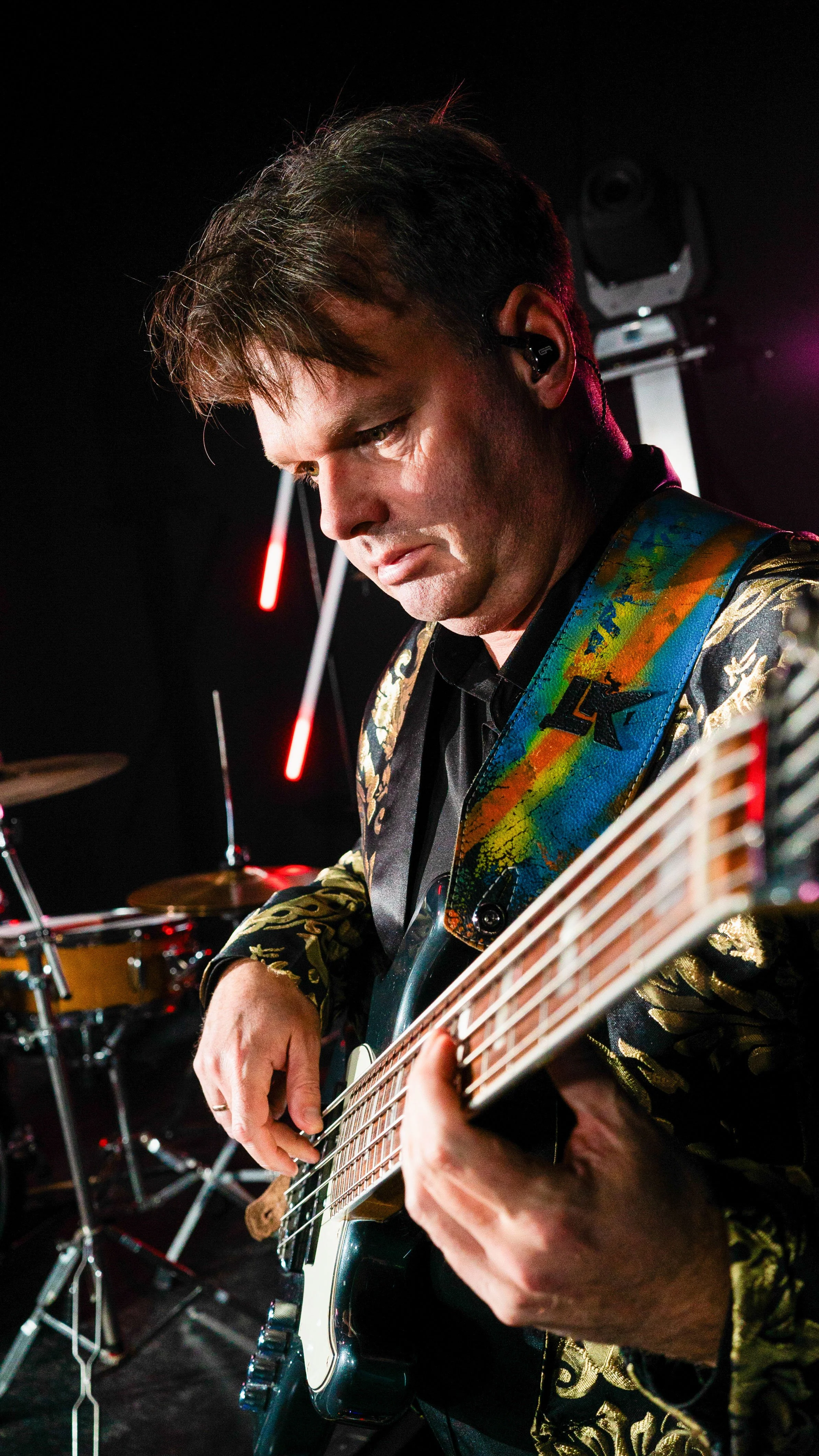A man playing an electric guitar on stage, wearing a colorful suit with gold details and an earpiece, with a drum set in the background.