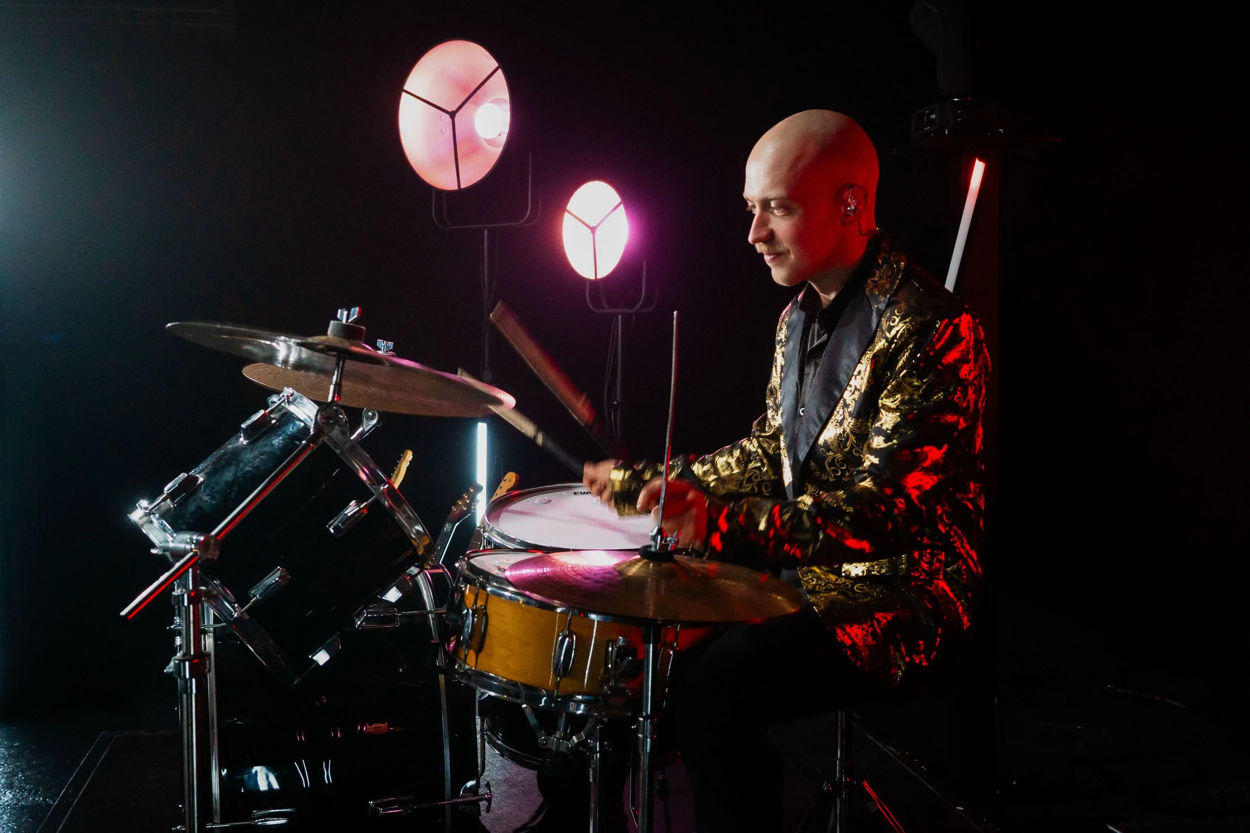 A man with a bald head, wearing a black and gold patterned jacket, playing a drum set in a dark room with colorful stage lights in the background.