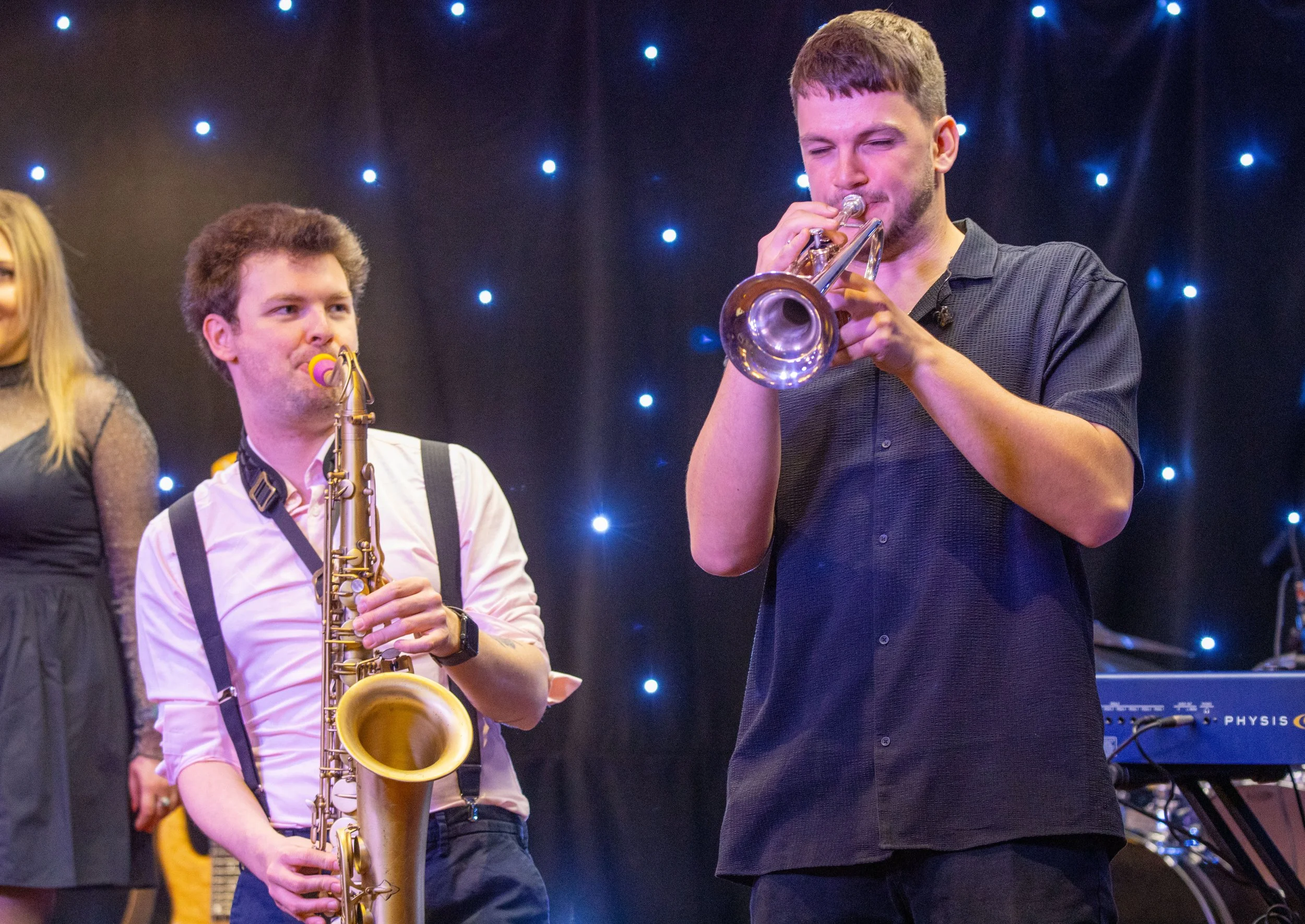A young man playing a trumpet and a young woman holding a saxophone on stage with a starry background of small lights.