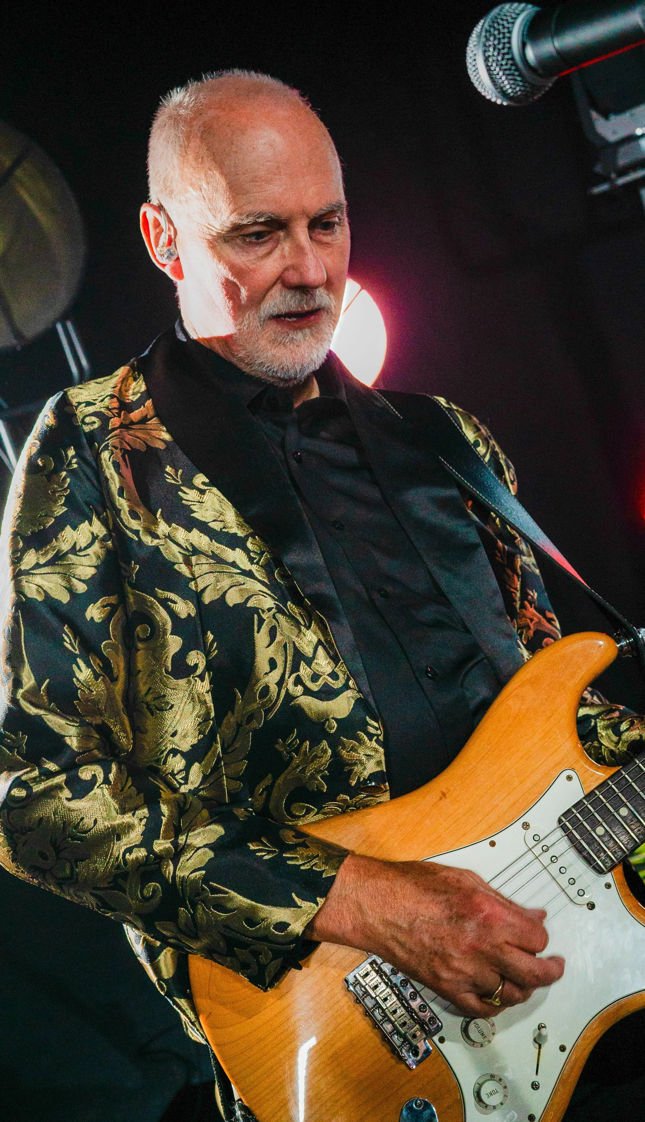 An older man with white hair and a beard playing an electric guitar on stage, wearing a black shirt and a gold patterned blazer, with a microphone visible to his right