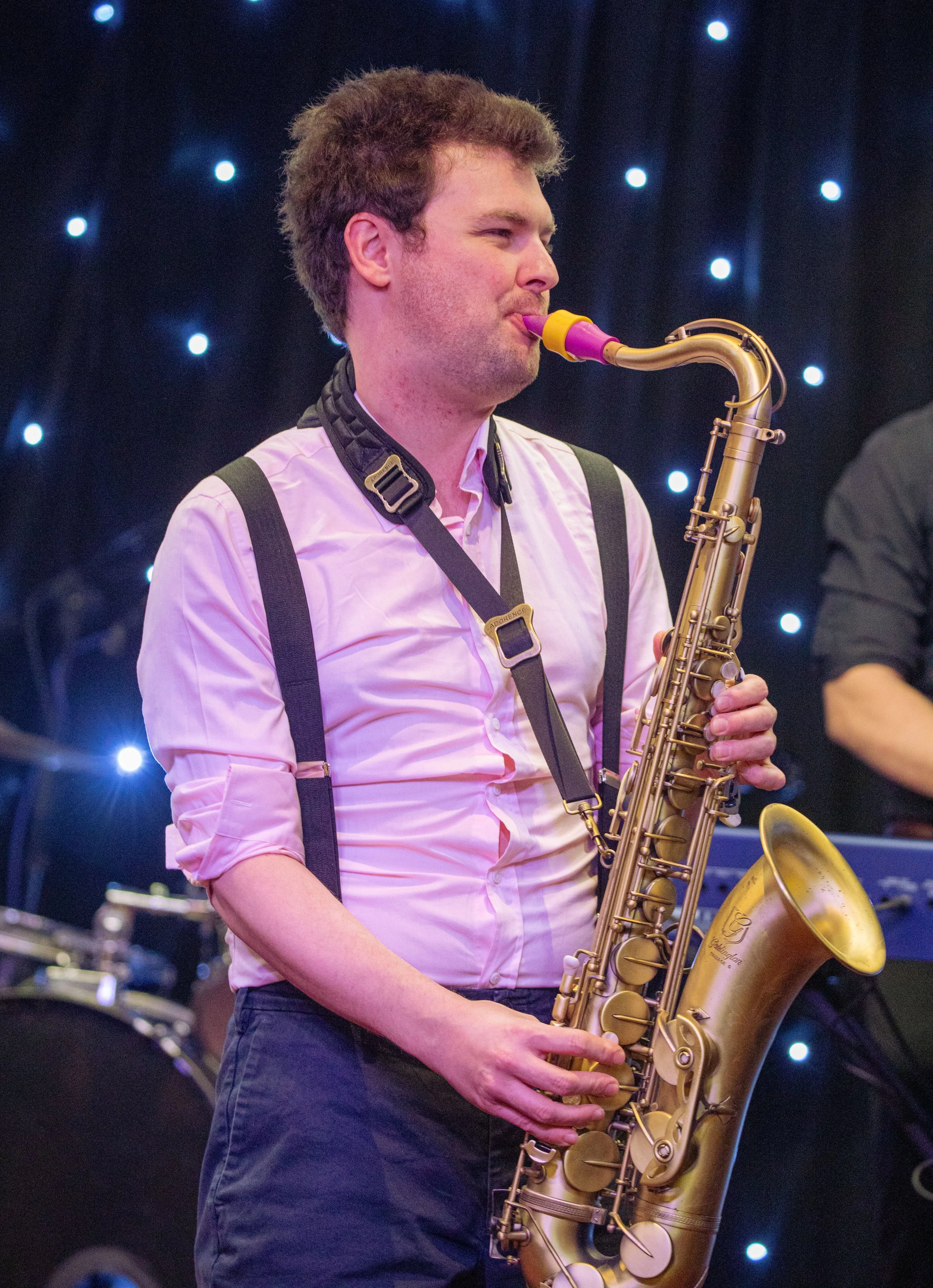 A man playing a gold saxophone at a musical performance with a dark background featuring small illuminated lights.