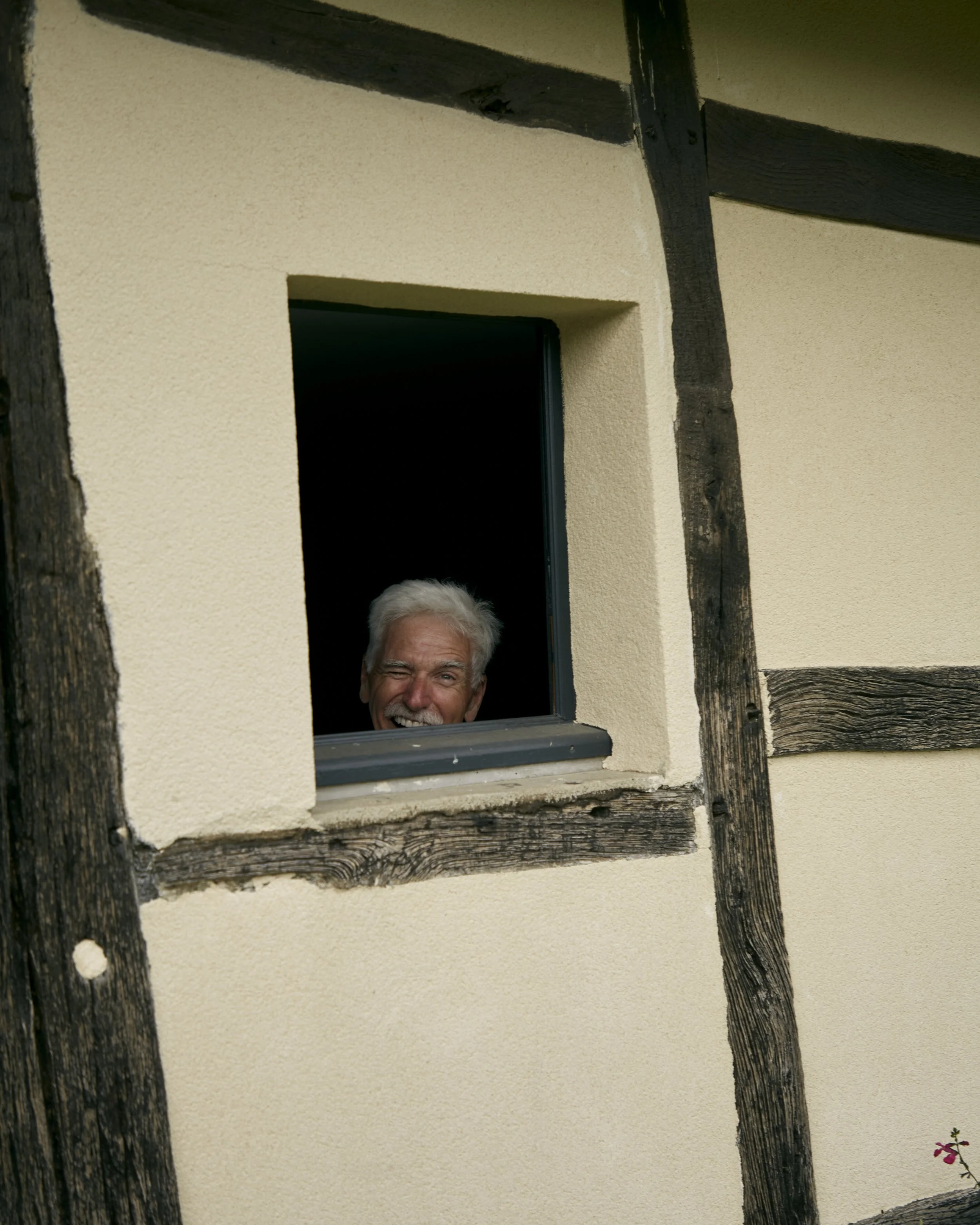 Photo d'une maison avec un mur beige, des poutres en bois noir, une fenêtre ouverte et une petite fleur à la base du mur.