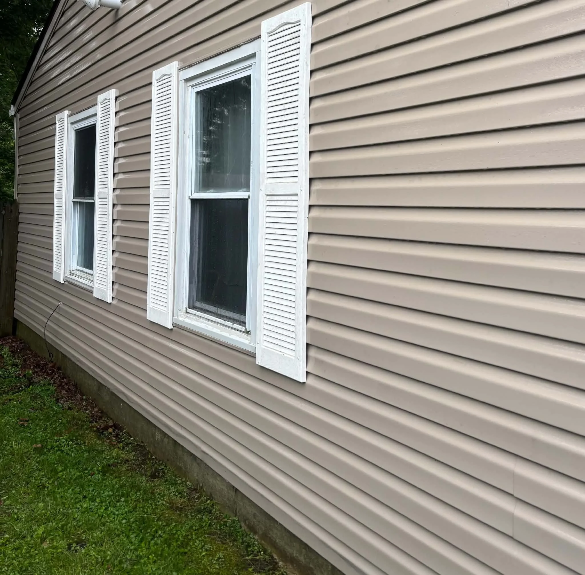 Two windows with white shutters on beige vinyl siding of a house. The house is situated on a grassy area with trees visible in the background.