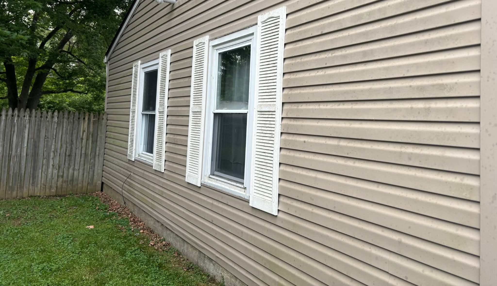 Exterior view of a house with beige vinyl siding, white window shutters, two windows, a wooden fence, green grass, and a tree in the background.