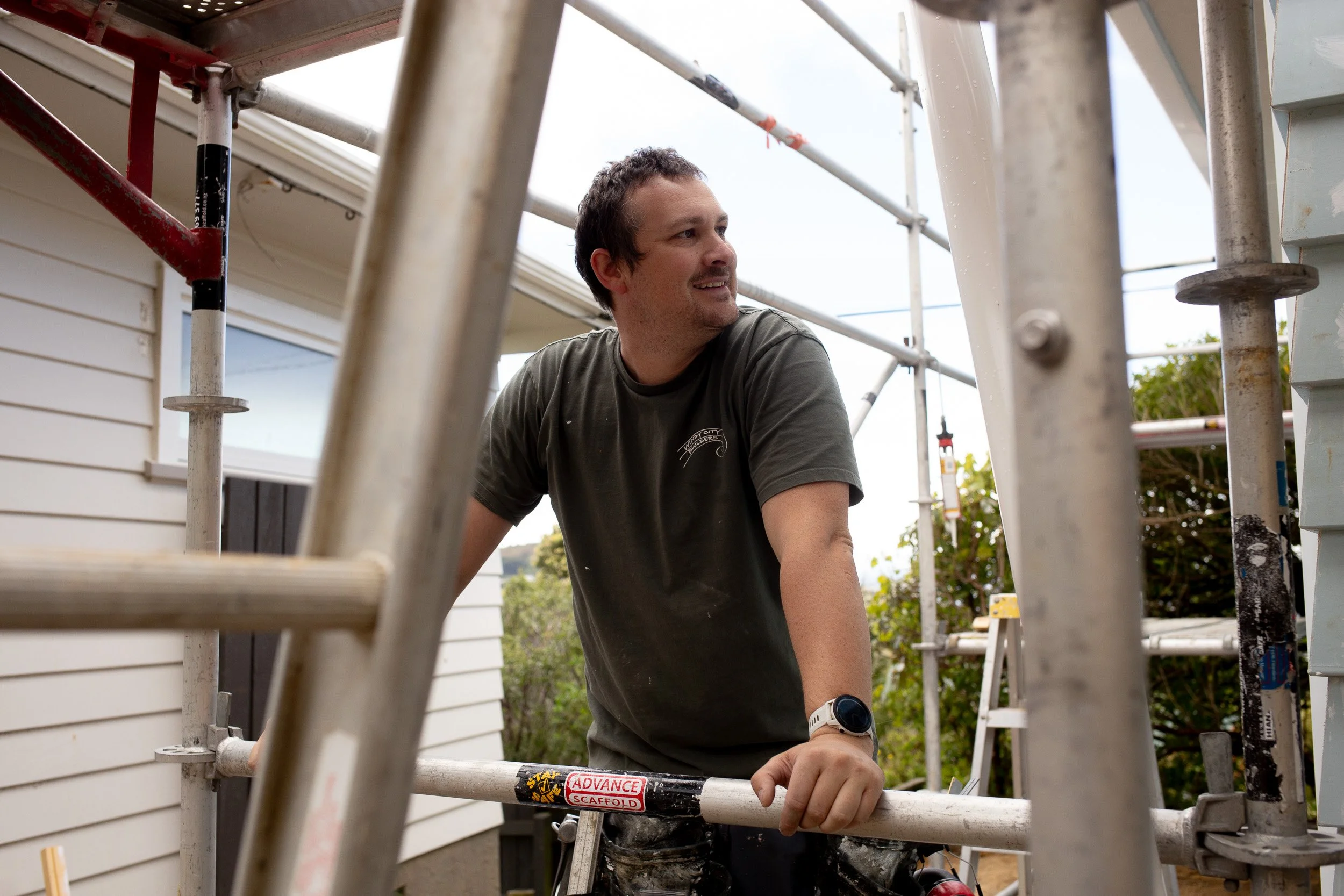 Alex, the founder of Windy City Builders, working on construction scaffolding outside a house, wearing a green shirt and black pants, with trees and a cloudy sky in the background.