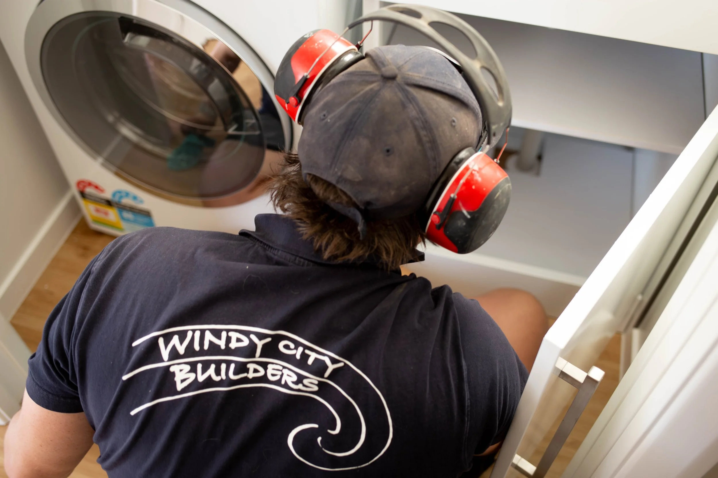 A person wearing earmuffs and a cap, leaning over an open front-loading washing machine in a laundry room.