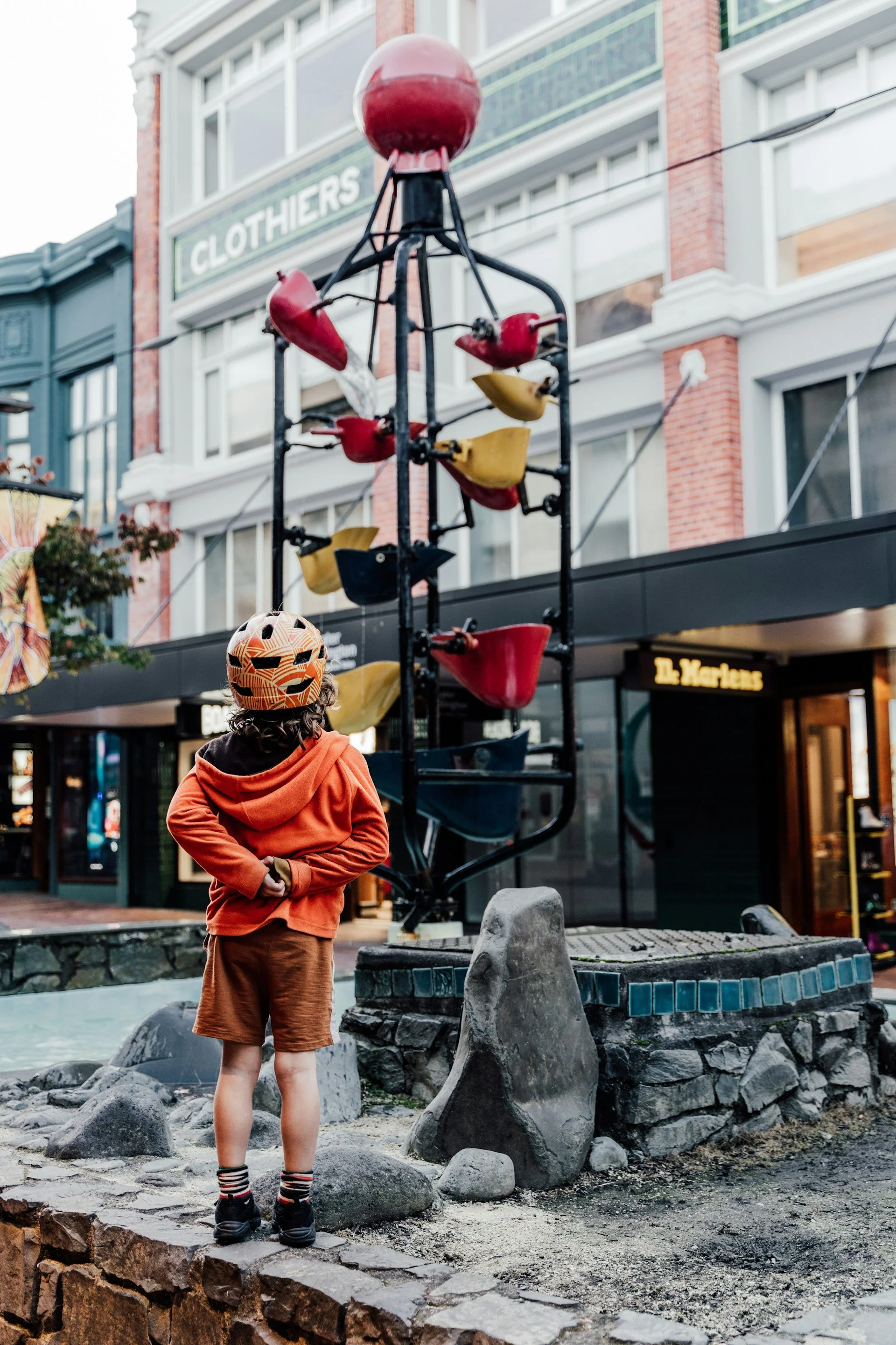 A young child with curly hair, wearing an orange hoodie, brown shorts, and striped socks, stands on a stone ledge and looks at a sculpture of a fish fountain in a city street. The fountain has colorful fish-shaped sculptures with water spouting from their mouths, set against buildings with storefronts, including one with a sign that says "CLOTHIERS."