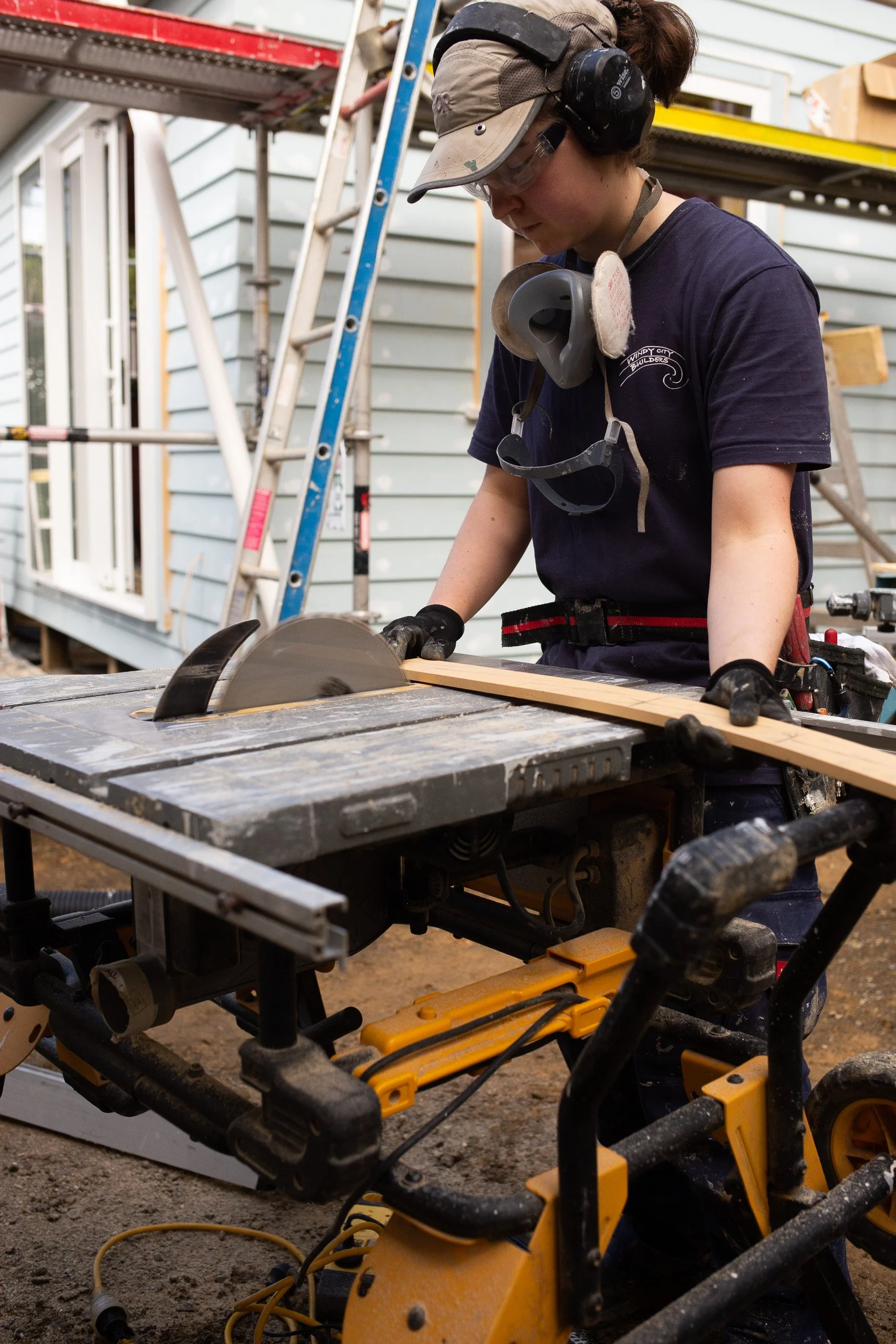 A person working on a construction site, operating a circular saw to cut wood. The person is wearing protective ear muffs, goggles, and gloves. There are ladders and construction materials visible in the background.