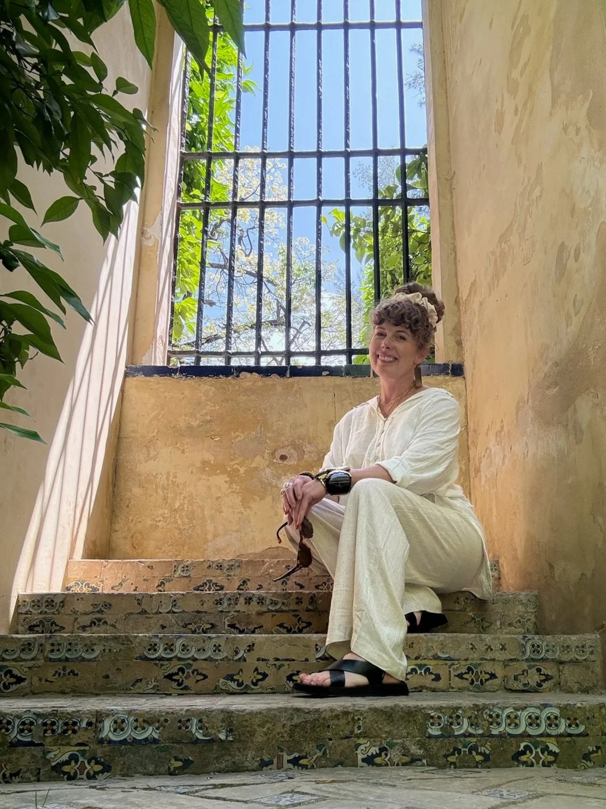 Portrait of writer Kara Dyas sitting on tiled stone steps beneath a sunlit iron gate.