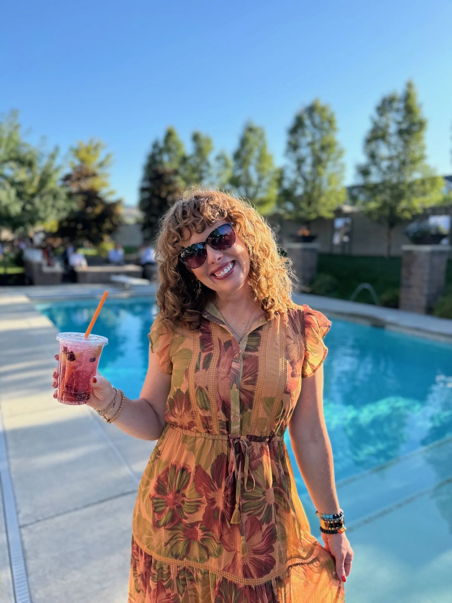 Kara is standing next to a pool in a long summer dress drinking a specialty seltzer