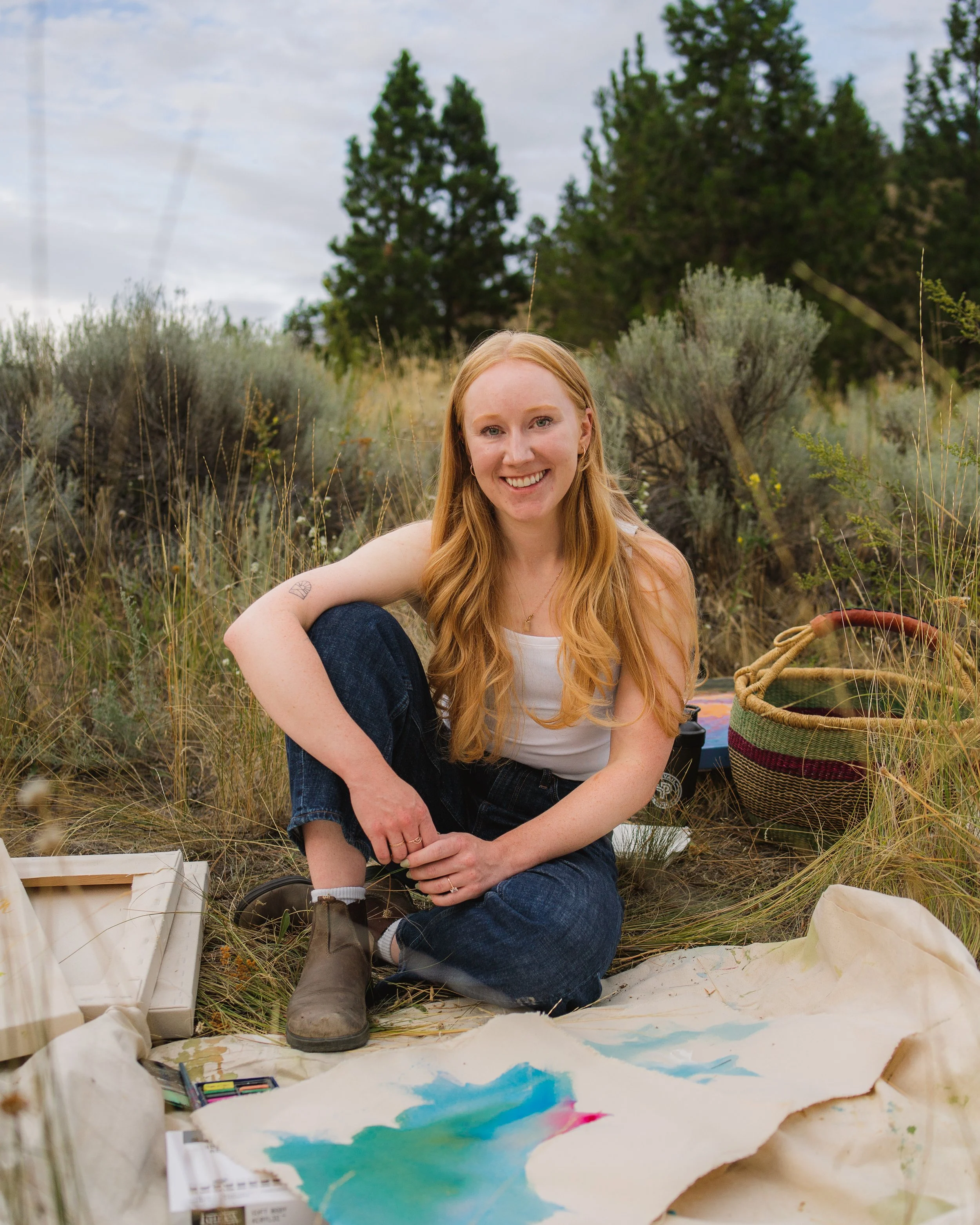 A woman with long red hair smiling while sitting outdoors on grassy ground with art supplies around her, including watercolor paints and a large sheet of paper with colorful painted designs. She is surrounded by trees and shrubs on a cloudy day.