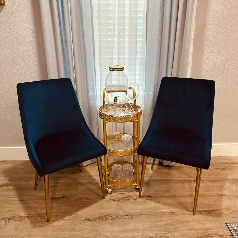 Two navy velvet chairs with gold legs flanking a gold rolling bar cart with glassware and a glass beverage dispenser, in front of a window at Avila Skin Studio in McKinney.