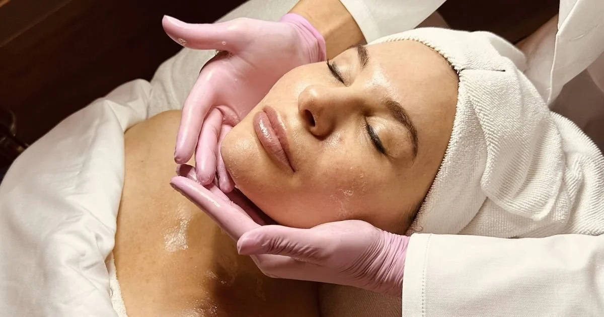 A woman receiving a facial treatment at Avila Skin Studio in McKinney, lying with her eyes closed, wearing a headband, while a professional in pink gloves applies or adjusts skincare on her face.