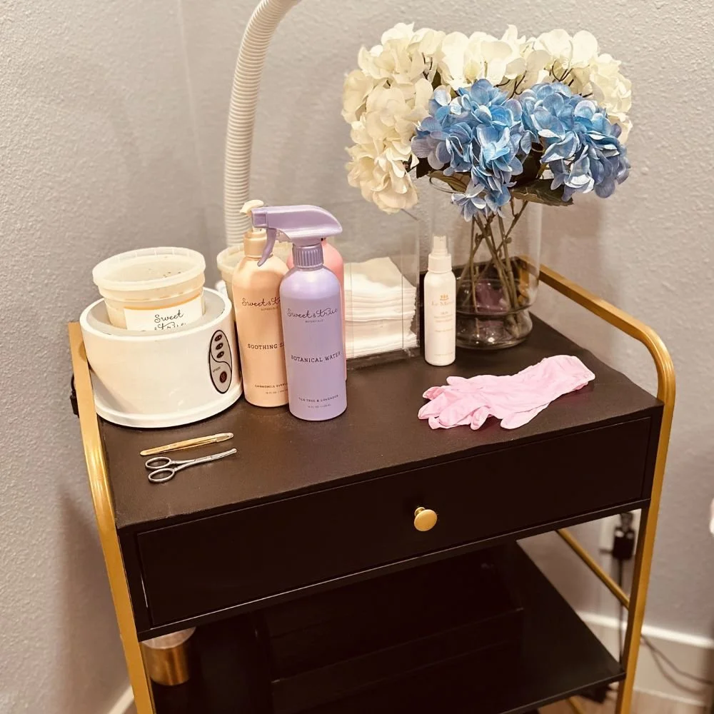A black and gold cart with a drawer and shelf, holding beauty and skincare products like sprays, bottles, a jar, and a white spray bottle. There are pink gloves on top and a clear vase with white and blue hydrangea flowers. Some tools and a tissue bo