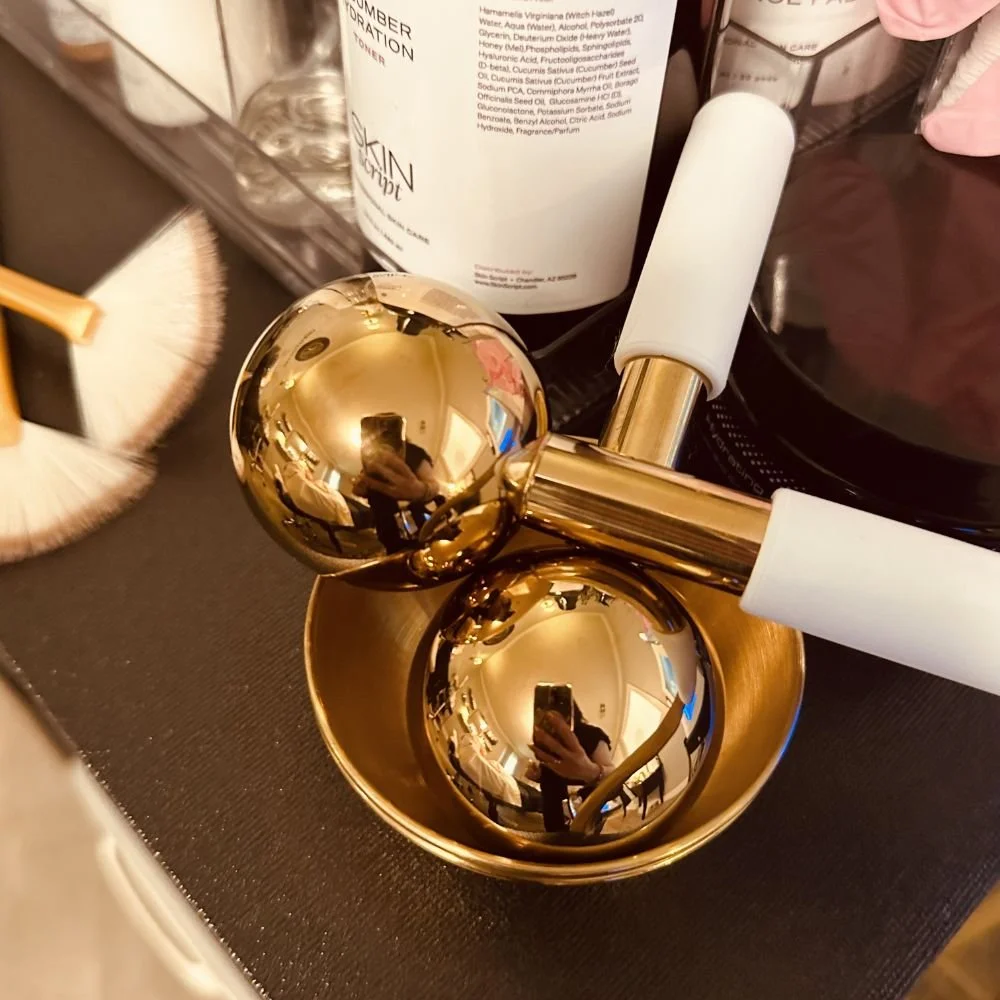 Close-up of a gold facial roller on a small gold dish, with reflection of a person taking a photo in the shiny surface. Surrounding items include a powder brush, bottles of skincare product, and a makeup brush.